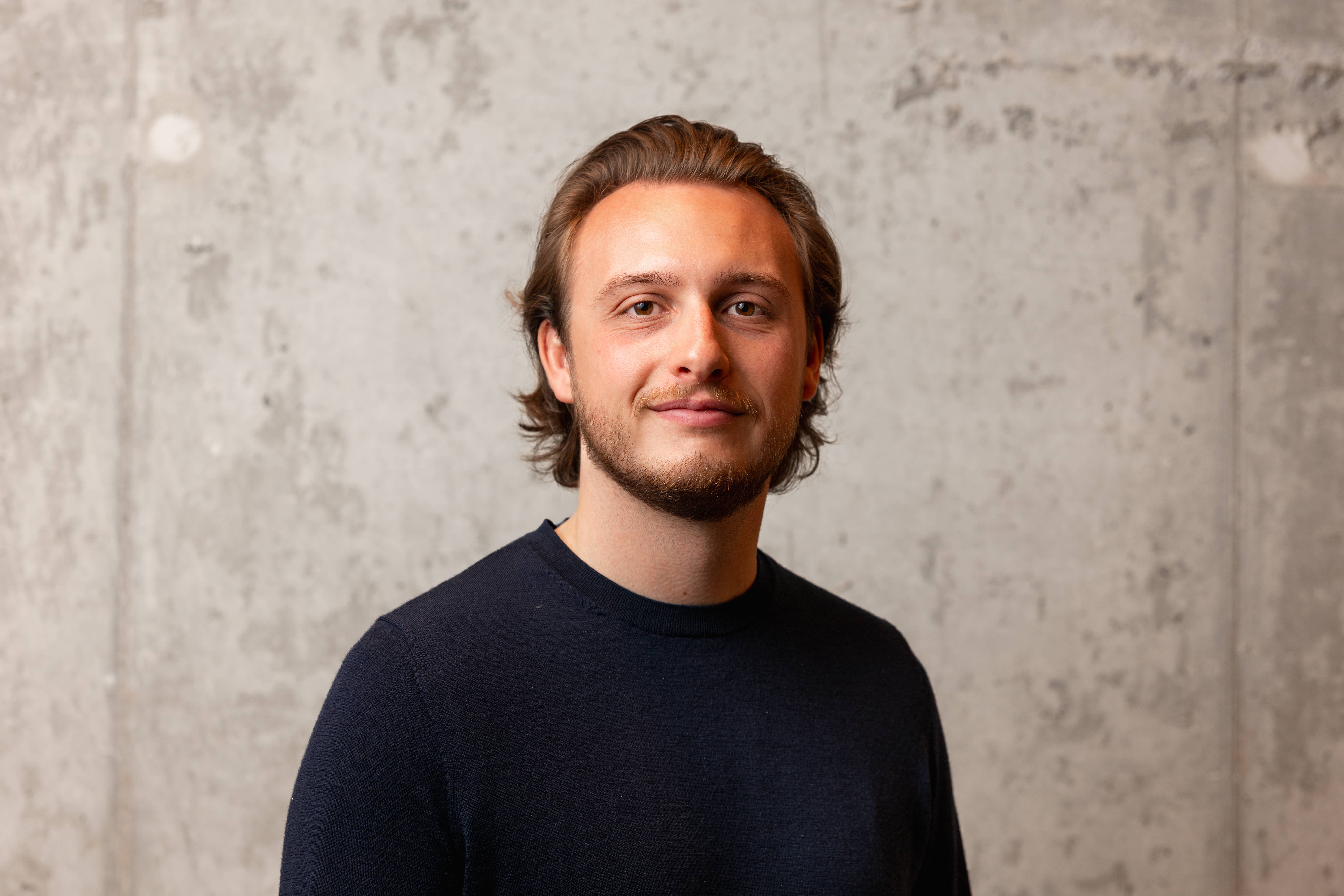 Young man with short brown hair and beard wearing a dark sweater standing against a textured gray wall.