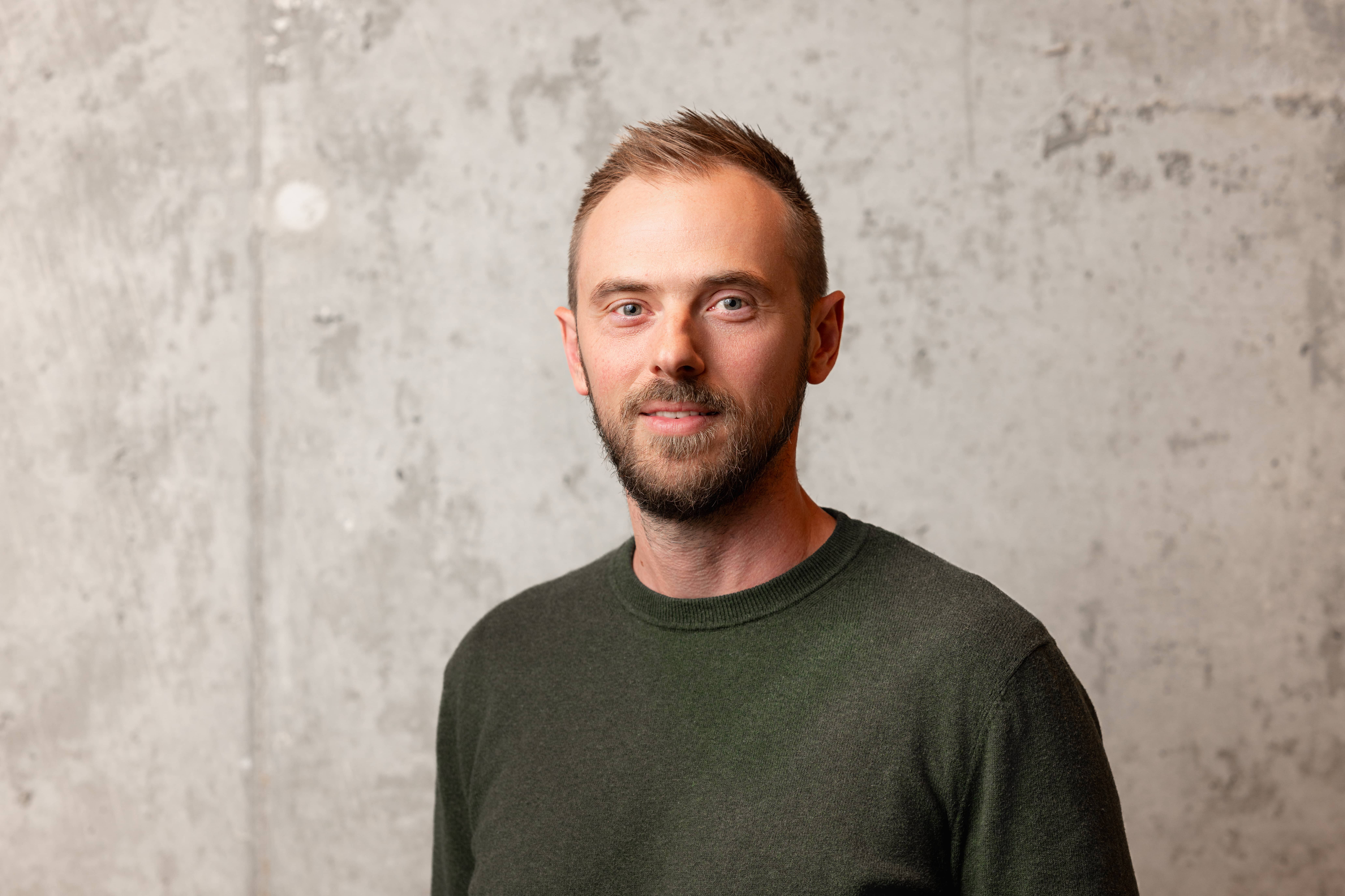 Portrait of a man with short brown hair and beard wearing a dark green sweater, standing in front of a light gray concrete wall.