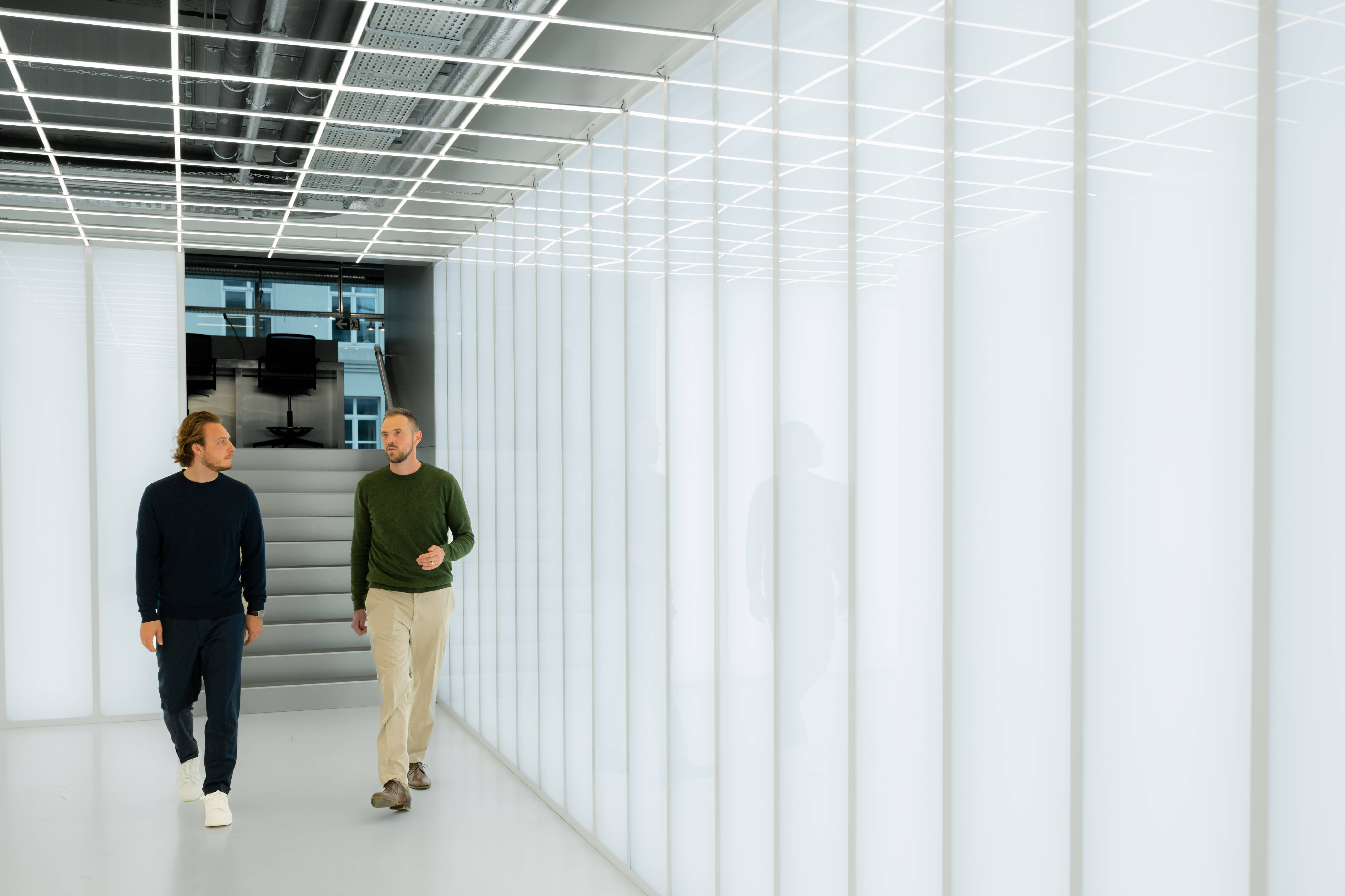 Two men walking and talking in a modern, brightly lit hallway with white walls and ceiling panels.