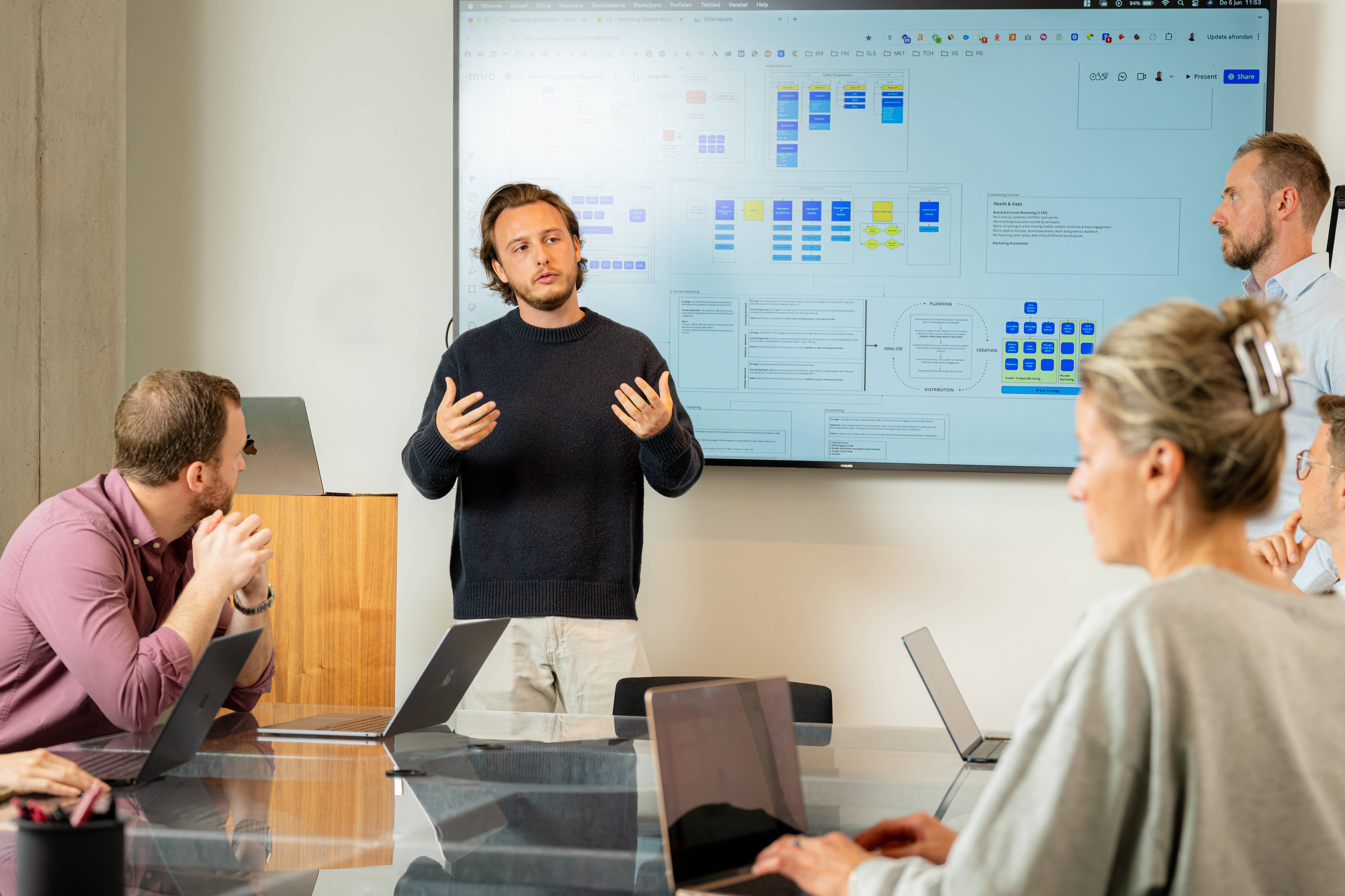 A man in a black sweater presenting to colleagues in a meeting room with a flowchart on a large screen behind him.