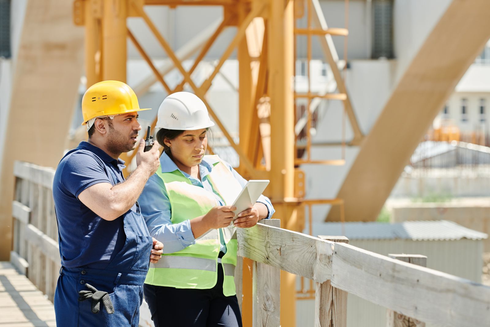 Two construction workers in hard hats on a job site, one using a radio while the other checks plans on a tablet