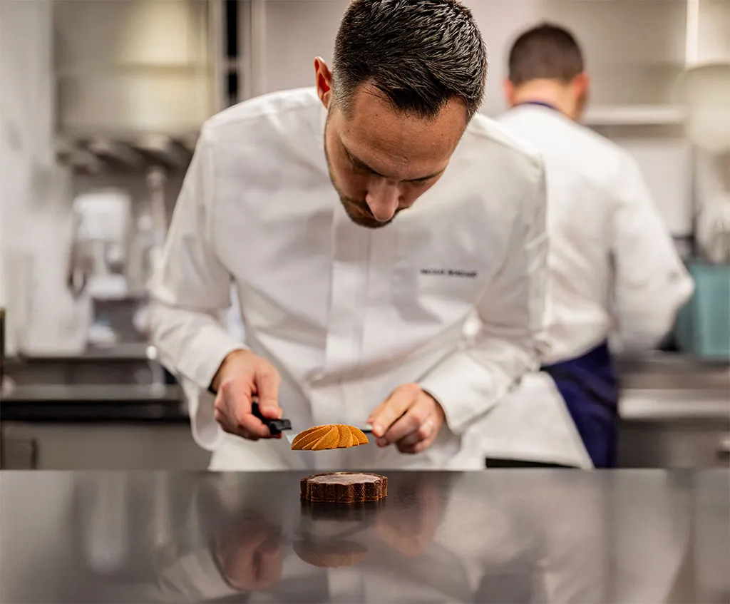 Nicolas Rouzaud in a white uniform, delicately place on the creation. The setting is a professional kitchen with another chef blurred in the background.