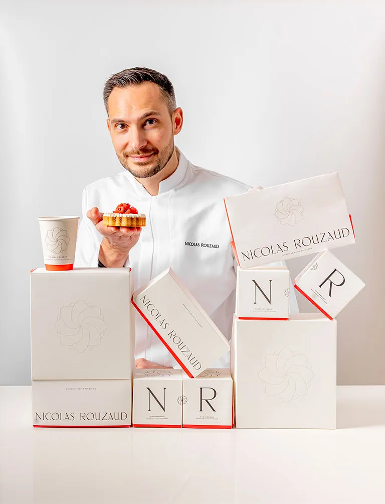 Nicolas Rouzaud smiles while sitting at a table with several white boxes from "Maison de Haute Patisserie." The boxes feature logos and the name "Nicolas Rouzaud." A coffee cup sits nearby, paired perfectly with a pastry crowned with a red topping.