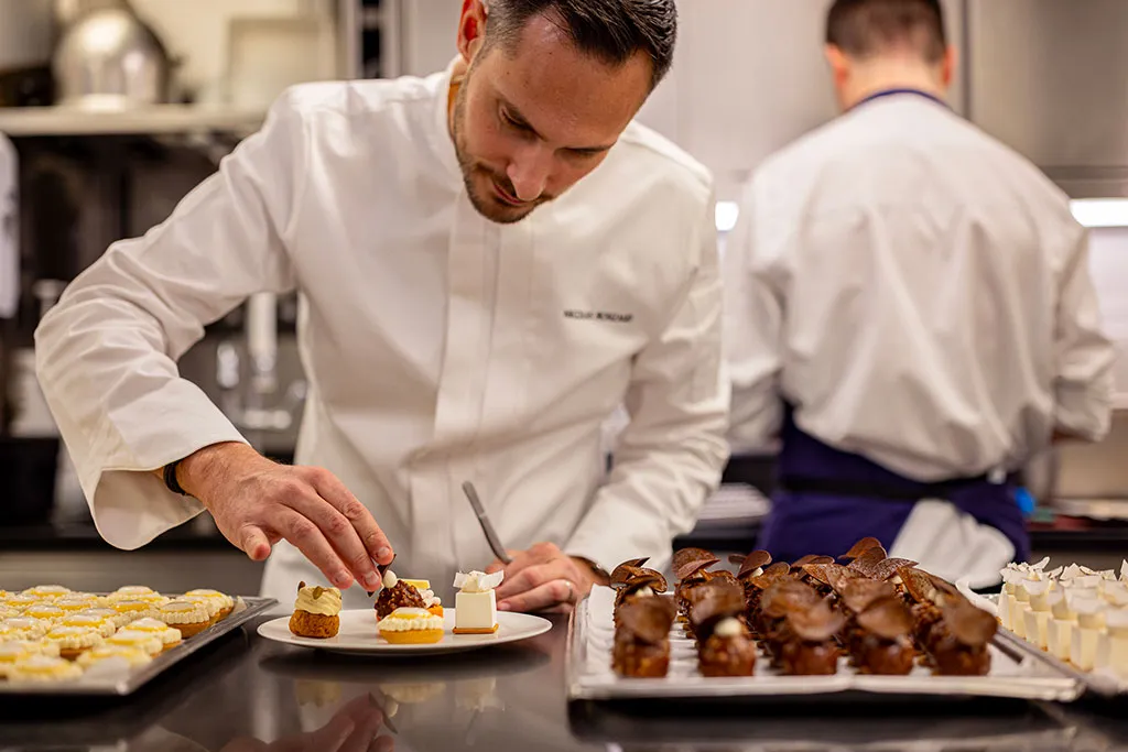 Nicolas Rouzaud in a white uniform, delicately place on the creation. The setting is a professional kitchen with another chef blurred in the background.