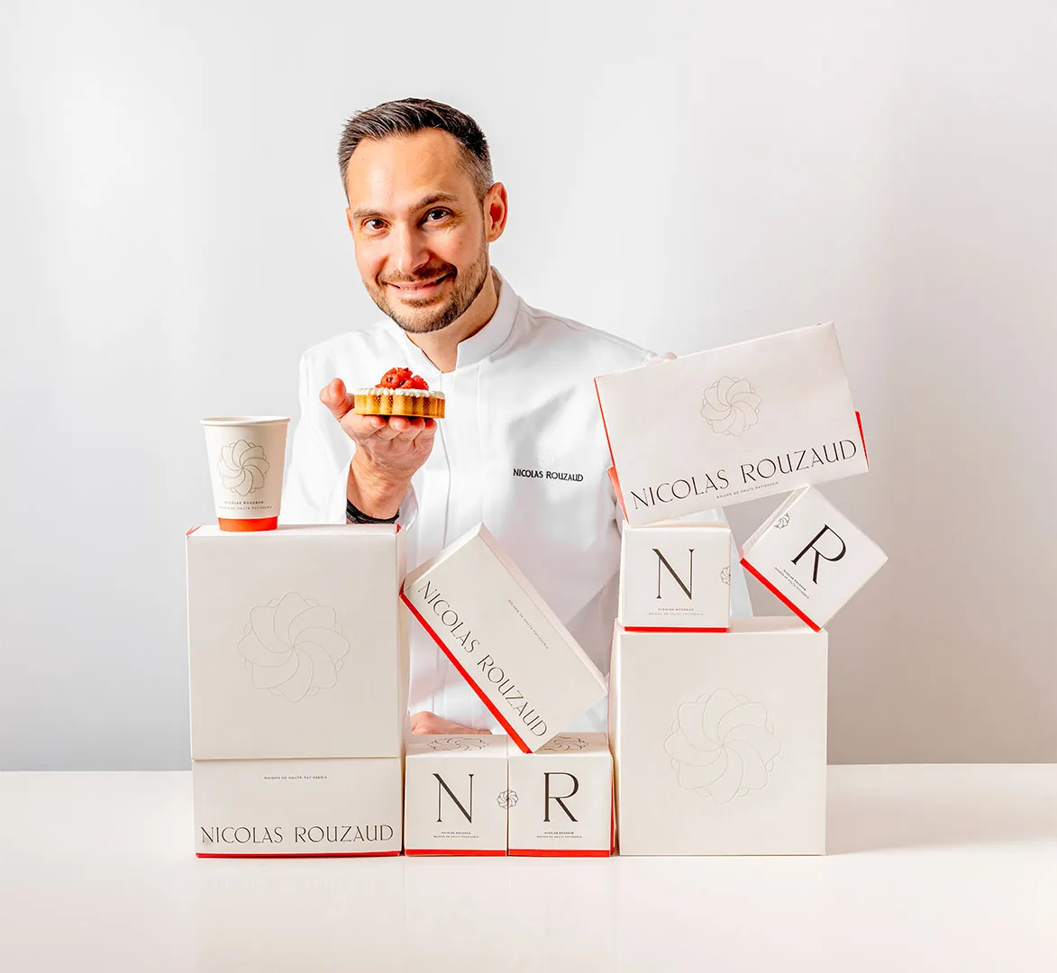 Nicolas Rouzaud smiles while sitting at a table with several white boxes from "Maison de Haute Patisserie." The boxes feature logos and the name "Nicolas Rouzaud." A coffee cup sits nearby, paired perfectly with a pastry crowned with a red topping.