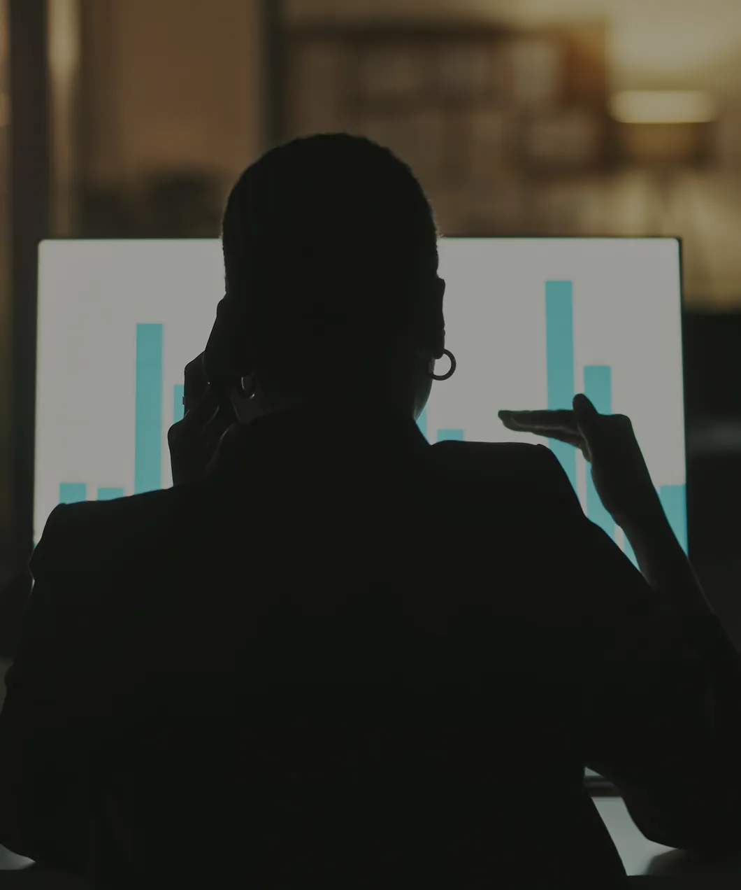 Silhouette of a person talking on the phone while pointing at a computer screen displaying a bar chart.