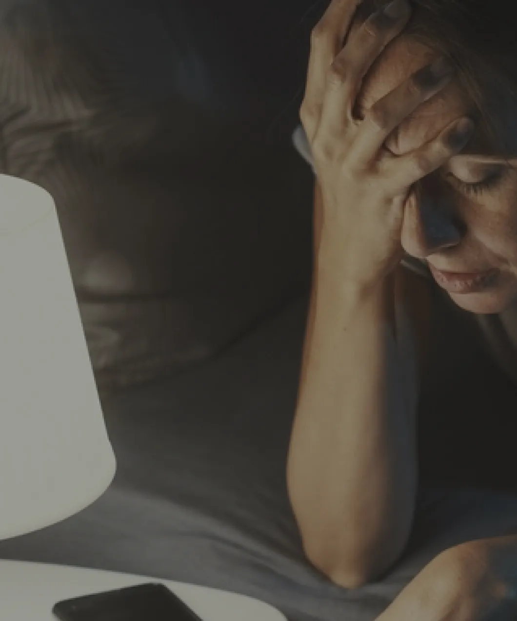Woman sitting in bed with eyes closed and hand on forehead beside a lit bedside lamp and a smartphone.