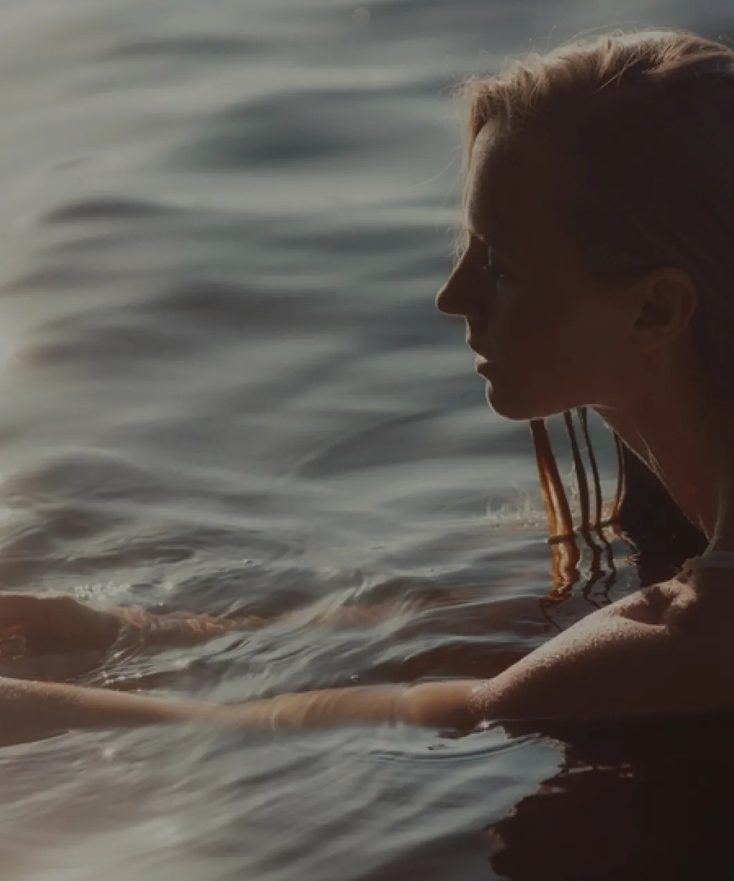 Side profile of a woman partially submerged in water with her arms extended forward, bathed in soft light.