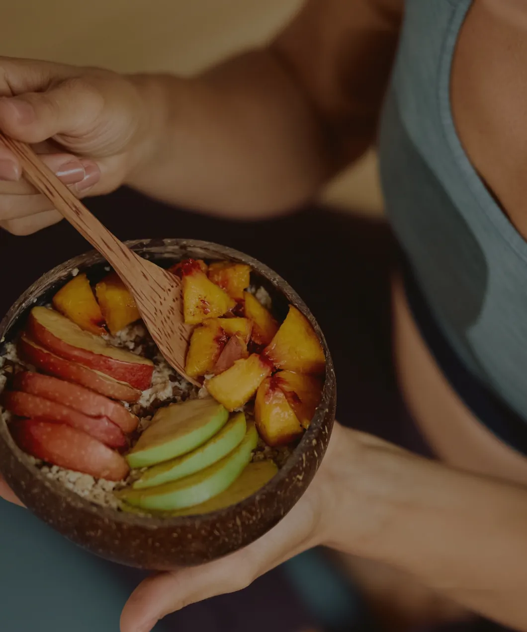 Person holding a wooden bowl with sliced peaches, apples, and green fruit, using a wooden spoon.