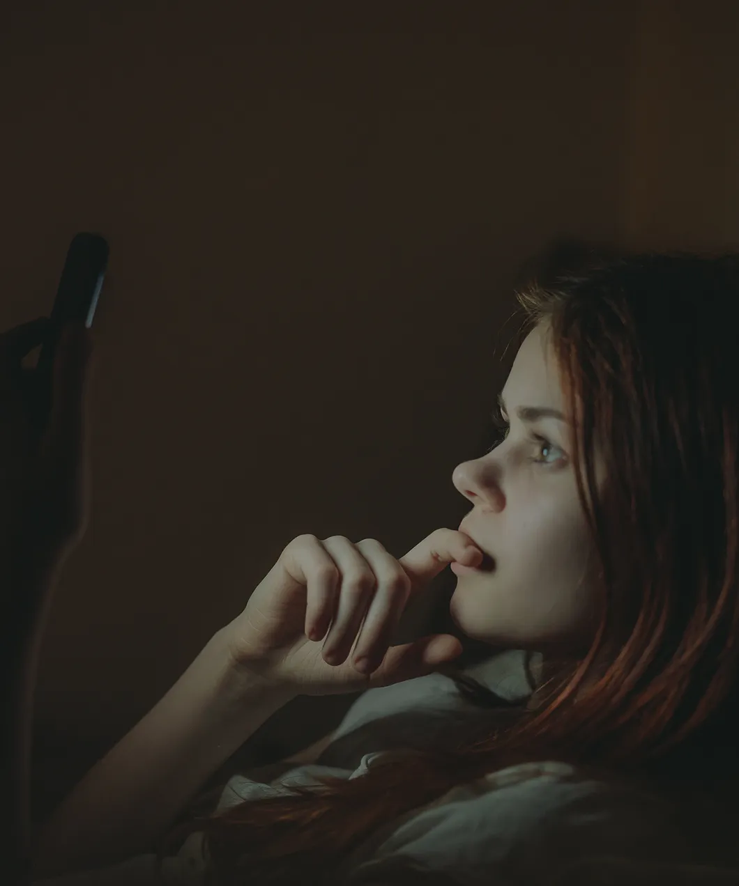 Young woman with red hair lying in bed at night, holding a phone and biting her finger anxiously.