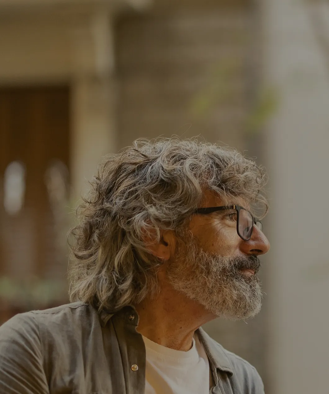 Side profile of an older man with gray curly hair, beard, and glasses looking thoughtfully to the right.