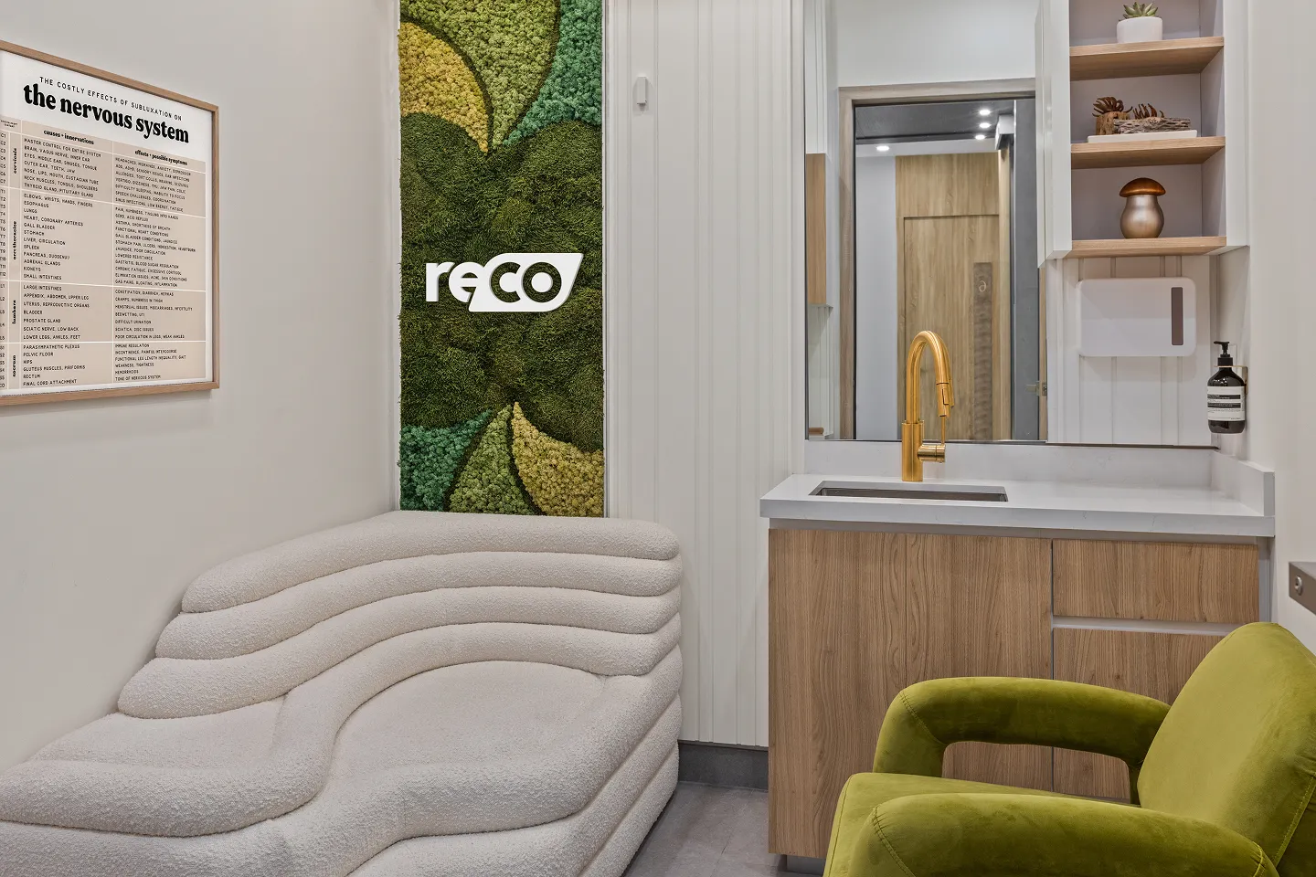 Modern clinic waiting area with a white textured sofa, green armchair, wooden sink cabinet with gold faucet, green moss wall art, and a framed nervous system chart.