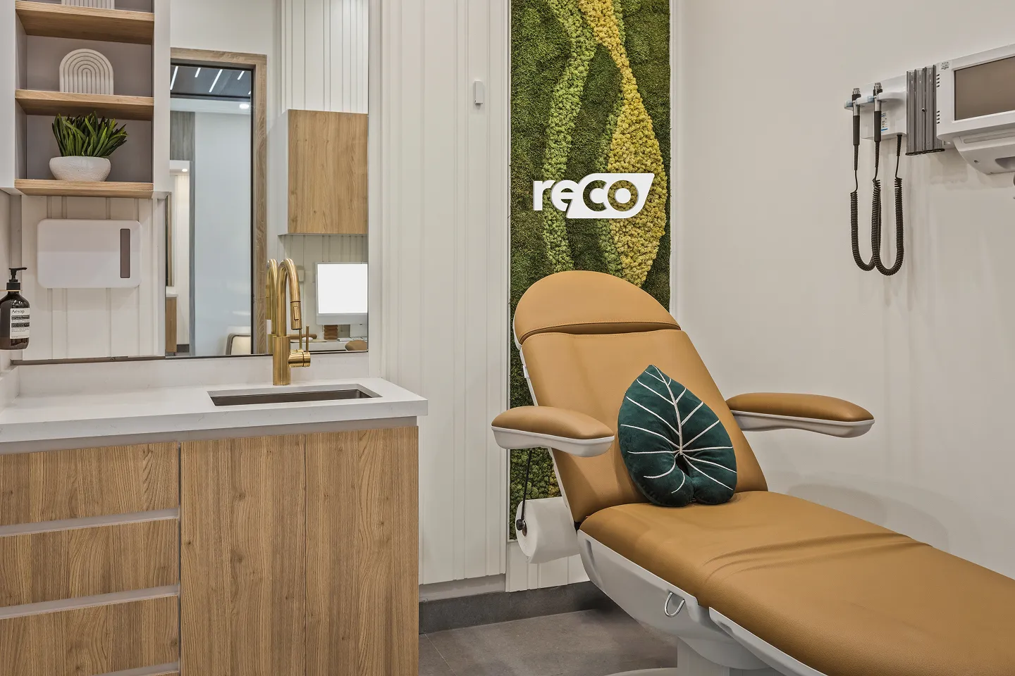 Modern medical examination room with a tan patient chair, green leaf-shaped pillow, gold faucet, and a textured green moss wall panel with the word 'reCO'.