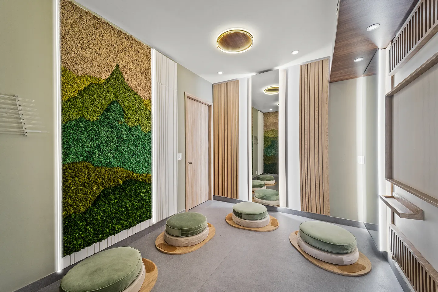 Modern room with green and brown moss wall art, circular green cushioned floor seats on wooden bases, vertical wood paneling, and a large mirror reflecting the ceiling light.
