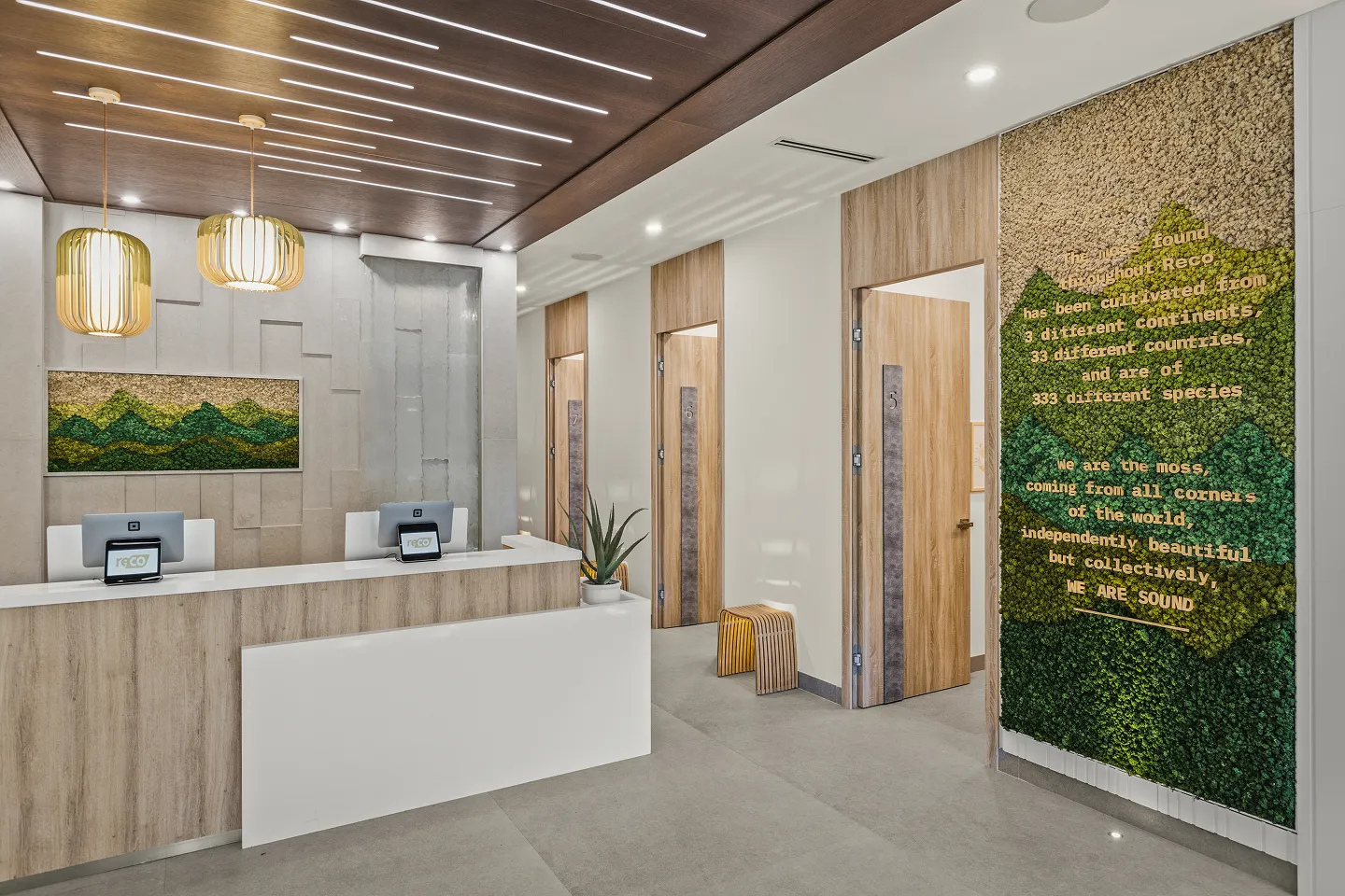 Modern reception area with wooden desk, two computers, two pendant lights, moss wall art behind desk and moss text wall near wooden doors.