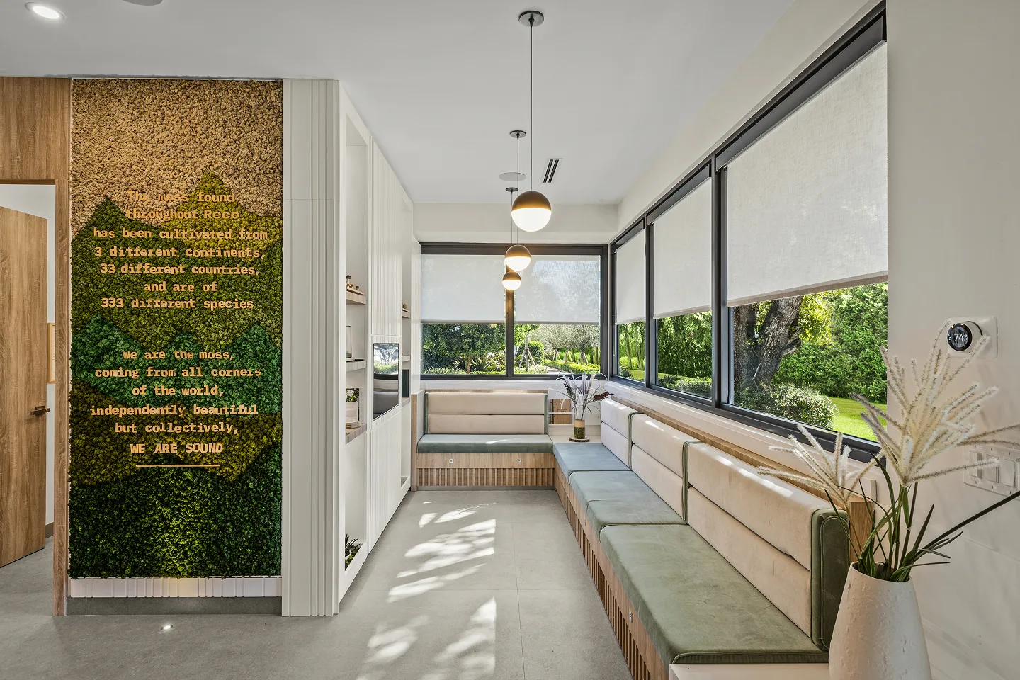 Modern waiting room with beige and green cushioned benches along large windows, pendant lights, and a decorative wall with green moss and gold text.
