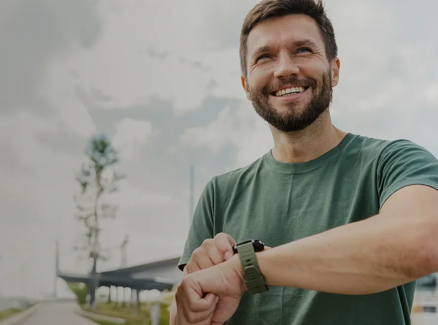 Smiling man in green t-shirt looking at his smartwatch outdoors with a cloudy sky background.