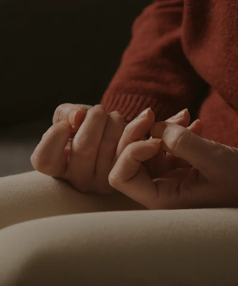 Close-up of a person’s hands resting on their lap, wearing a rust-colored knit sweater and beige pants.