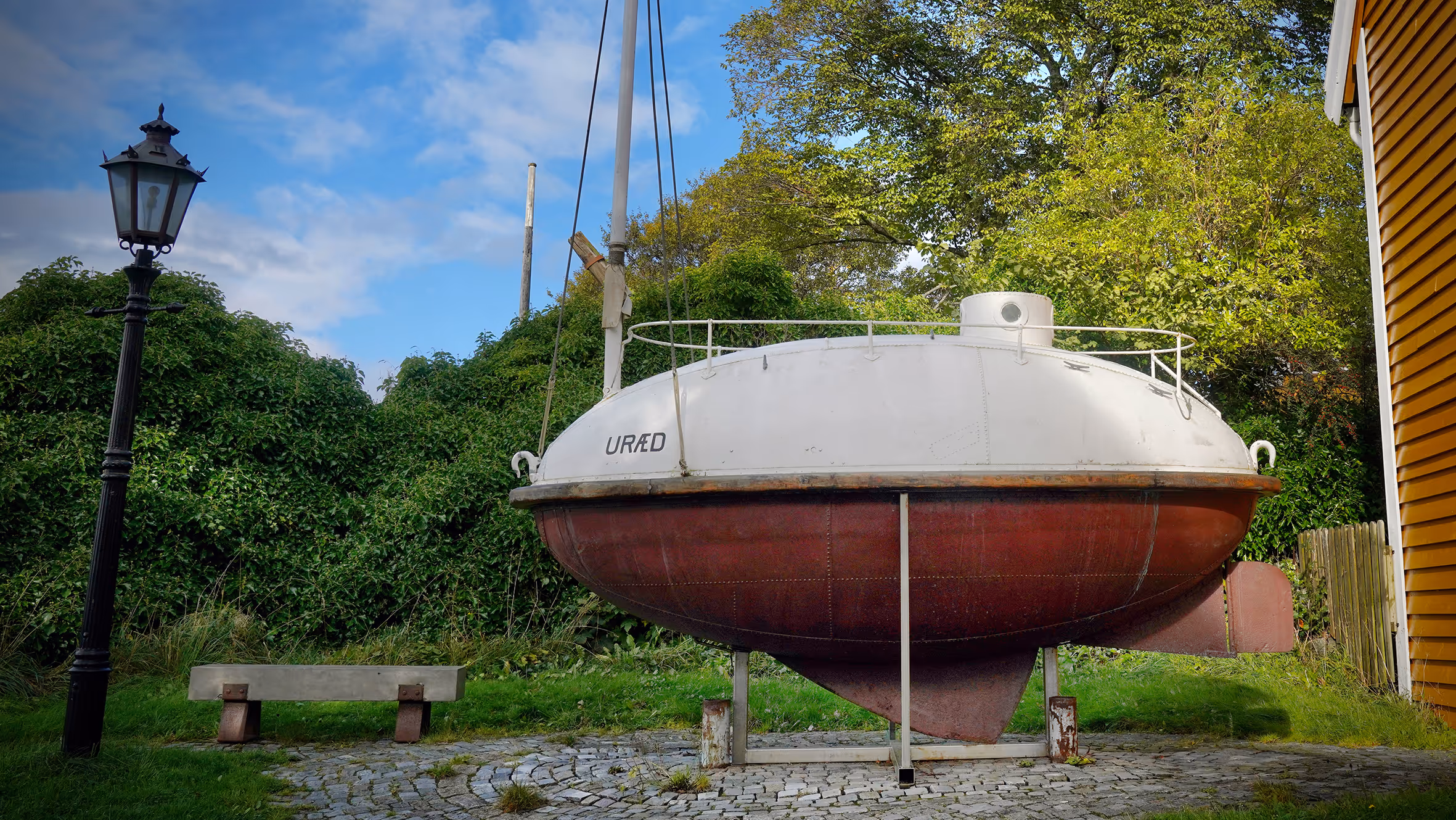 En rund ubåt med navnet URÆD, plassert på land ved siden av et gult hus, omgitt av grønn vegetasjon og en svart gatelykt.
