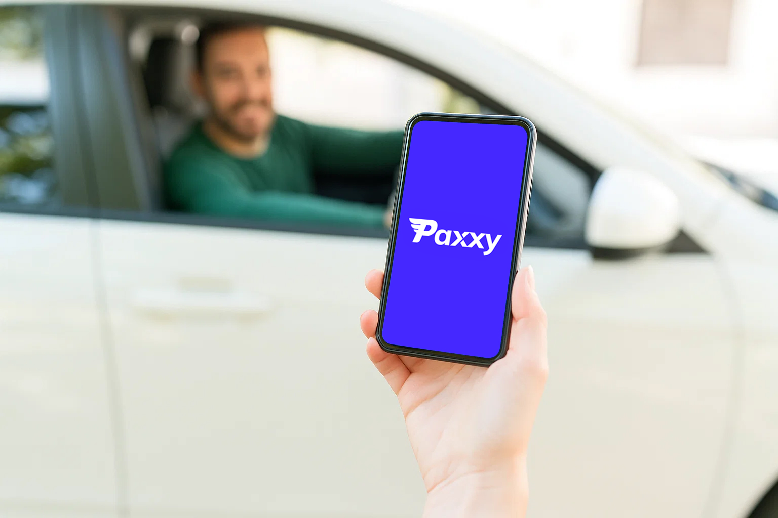 Hand holding a smartphone displaying the Paxxy logo in front of a man sitting inside a white car.