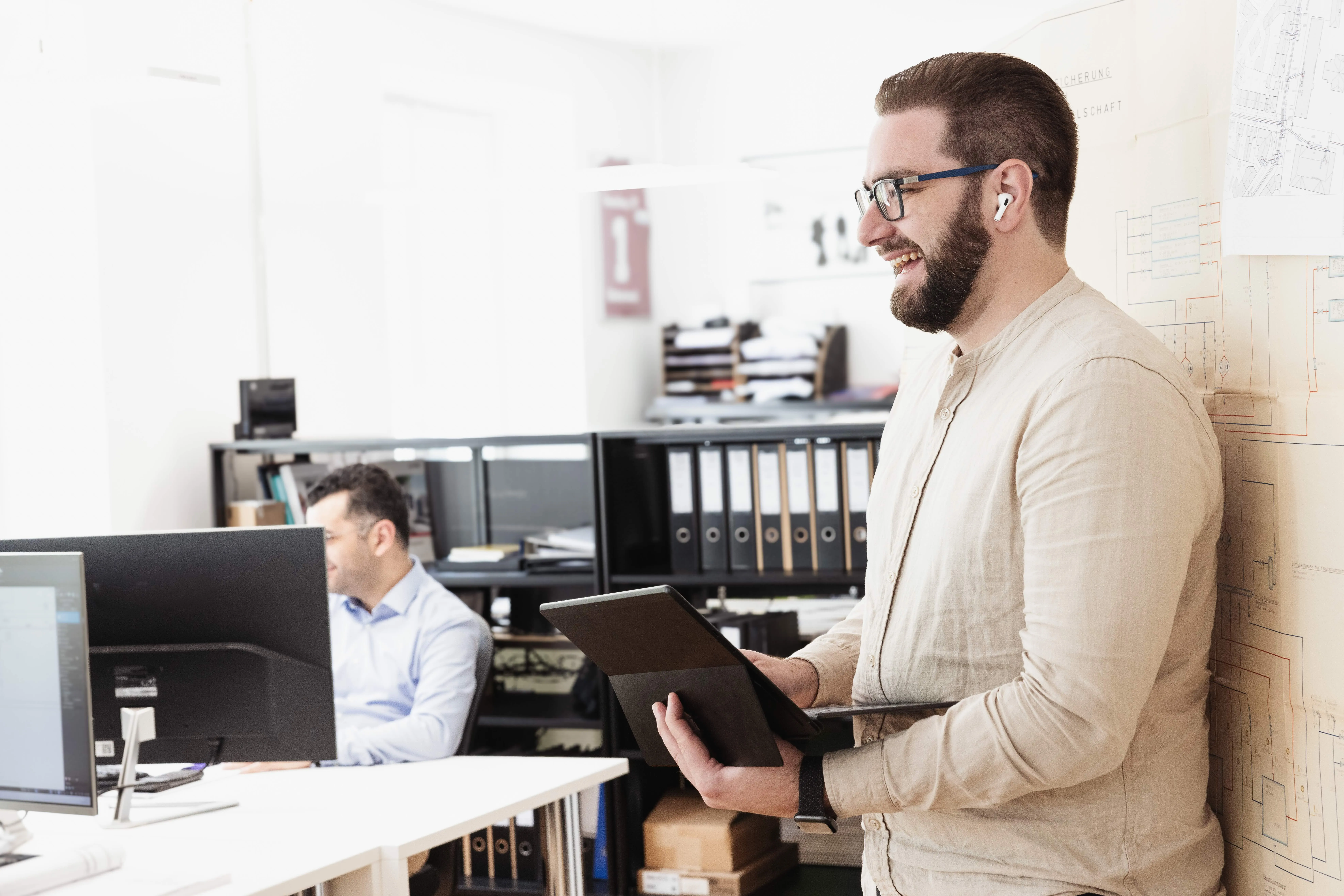 Mann mit Brille und Ohrhörern steht lächelnd mit einem Tablet in einem Büro, während im Hintergrund ein Kollege am Schreibtisch arbeitet.