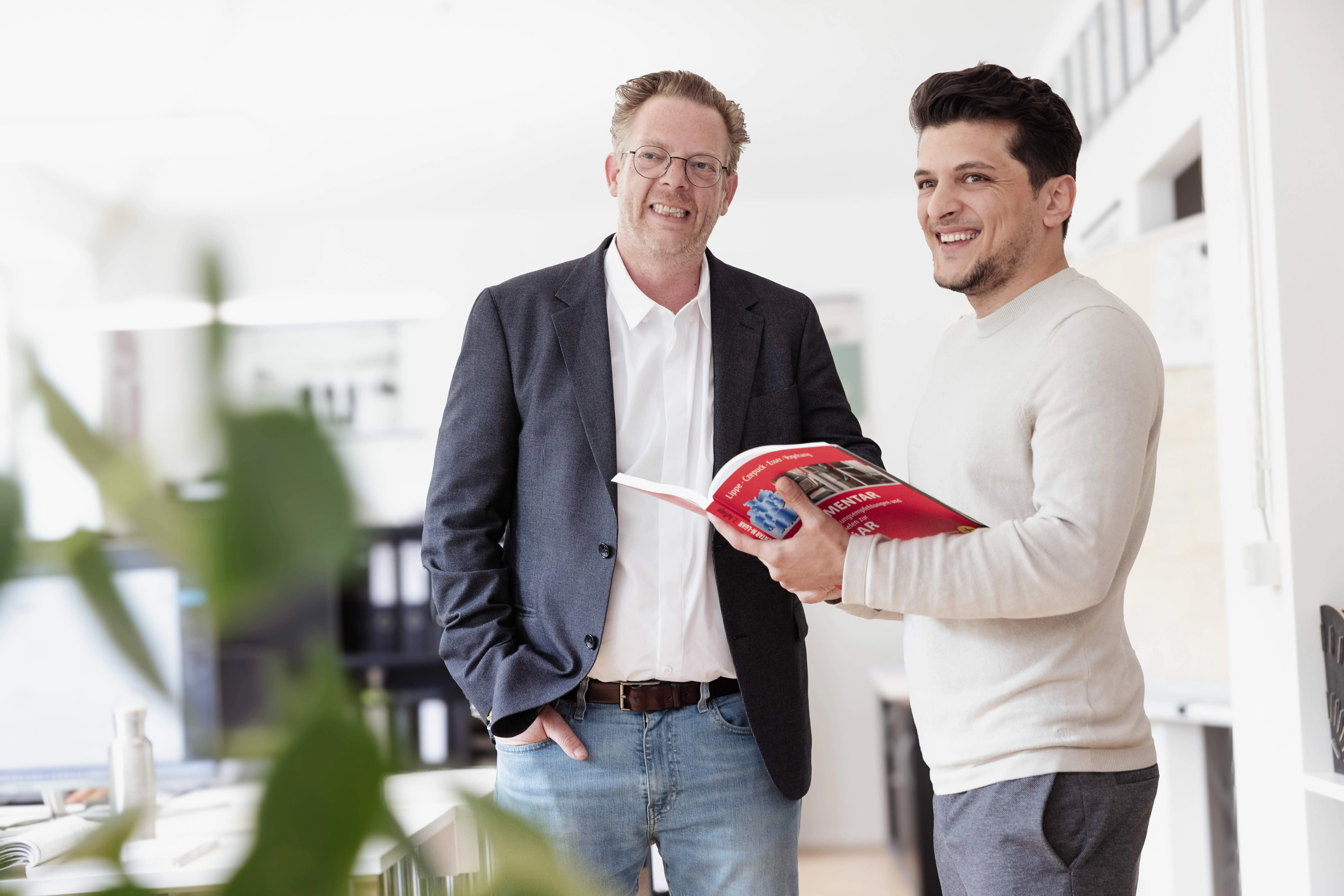 Zwei lächelnde Männer stehen in einem hellen Büro, einer hält ein rotes Buch.