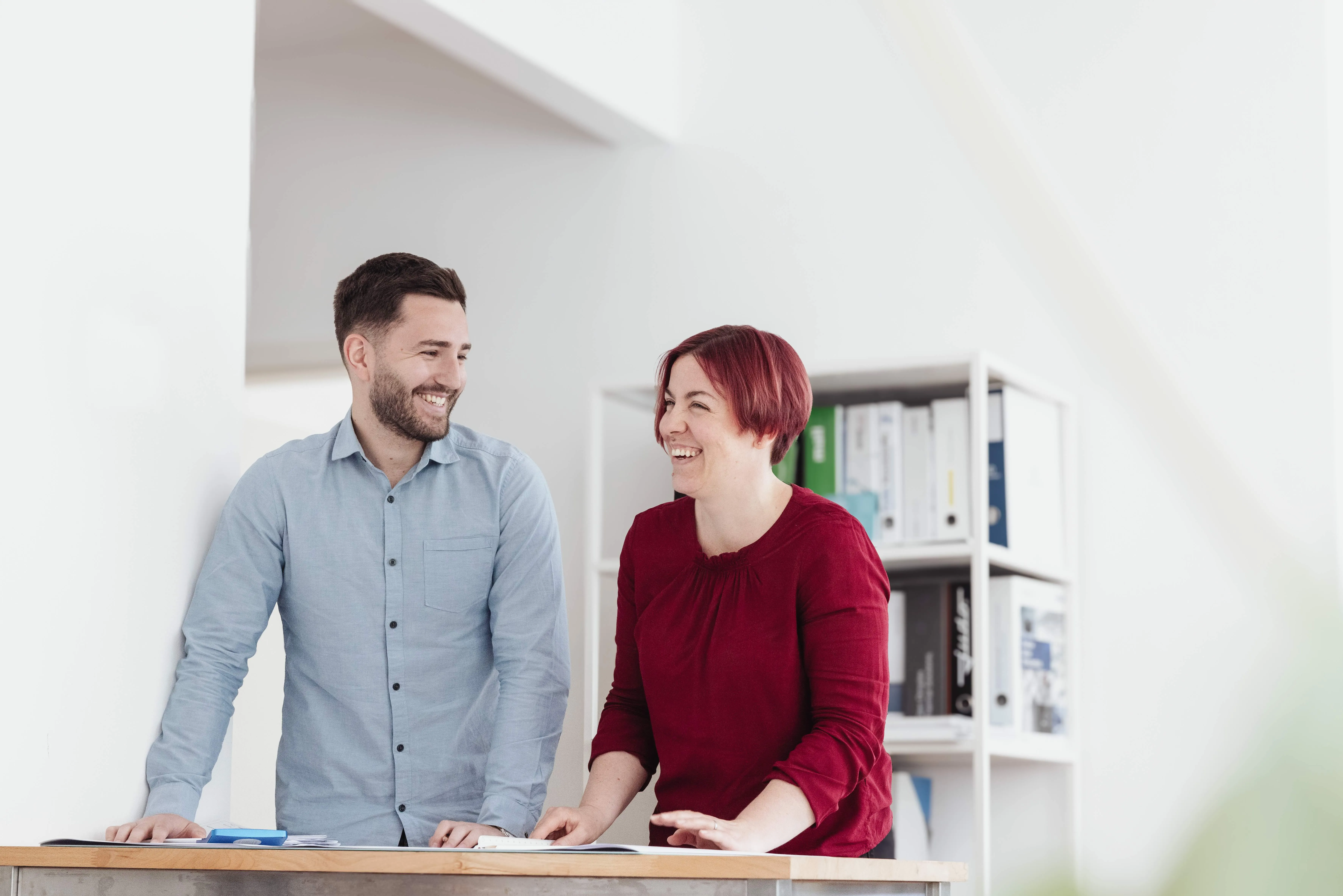 Zwei lächelnde Erwachsene, ein Mann im hellblauen Hemd und eine Frau in einem roten Pullover, die an einem Tisch stehen und sich unterhalten.