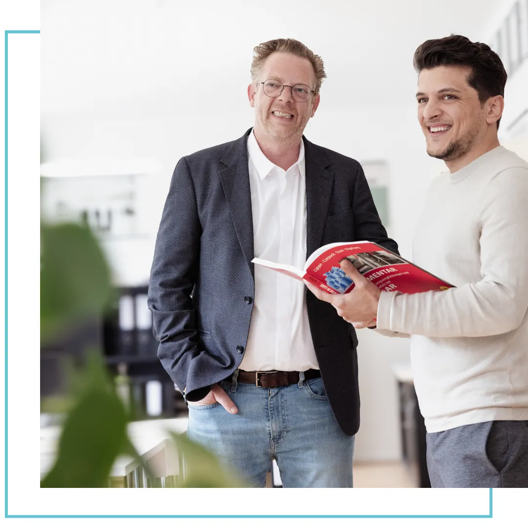 Zwei lächelnde Männer lesen gemeinsam ein rotes Buch in einem modernen Büro.