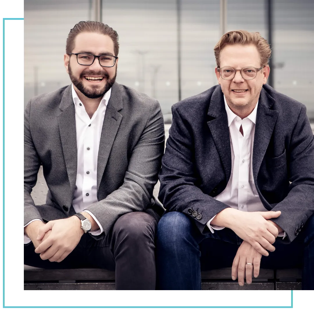 Zwei lächelnde Männer mit Brillen und grauen Blazern sitzen nebeneinander auf einer Bank.
