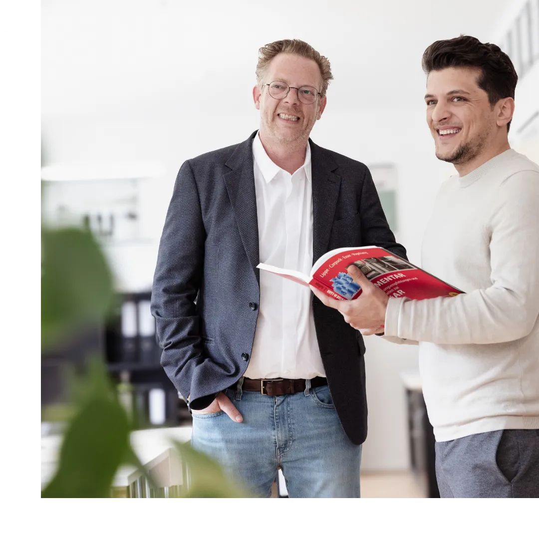Zwei lächelnde Männer stehen in einem hellen Büro, einer hält ein rotes Buch.