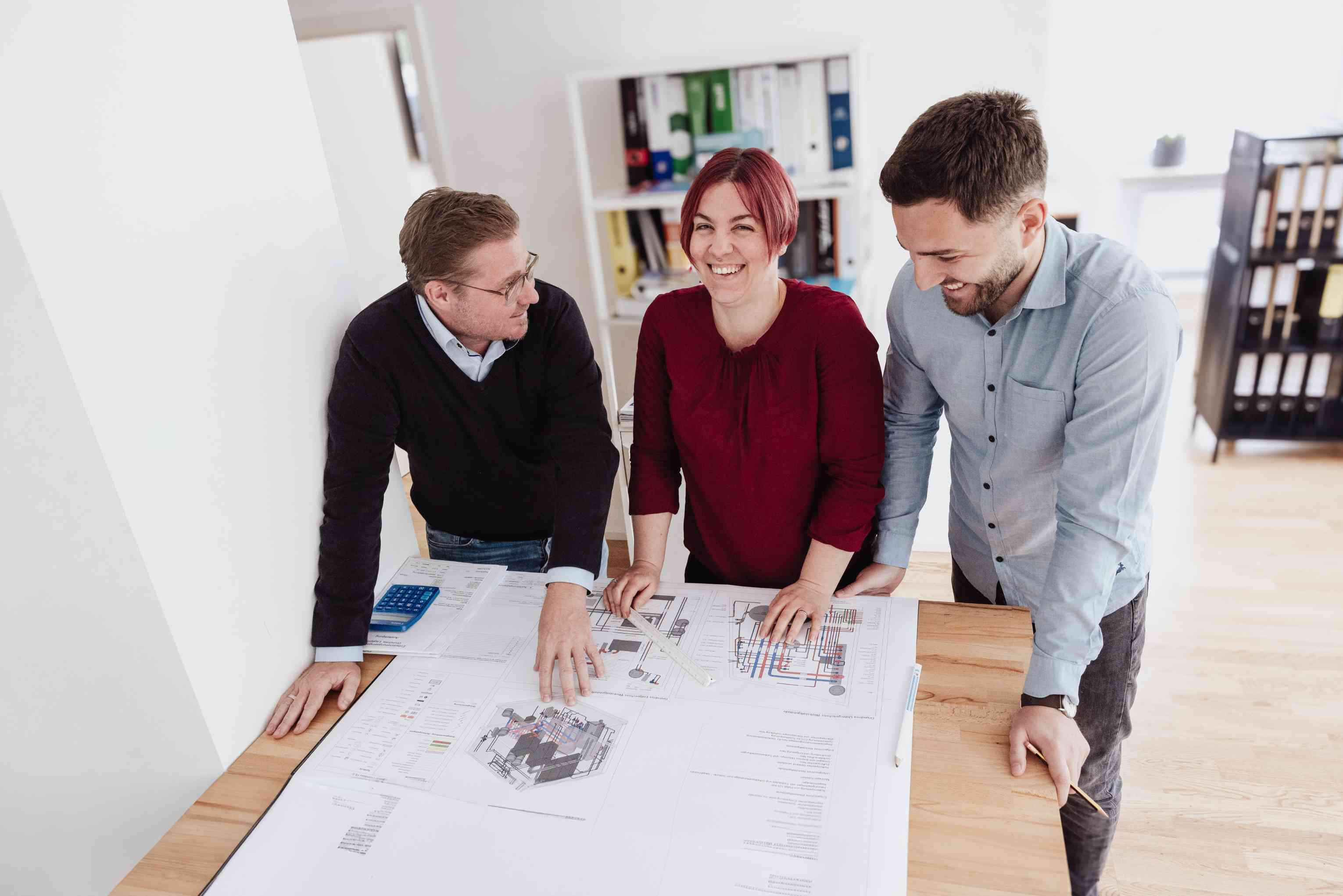 Drei Personen besprechen Baupläne an einem Holztisch in einem modernen Büro.
