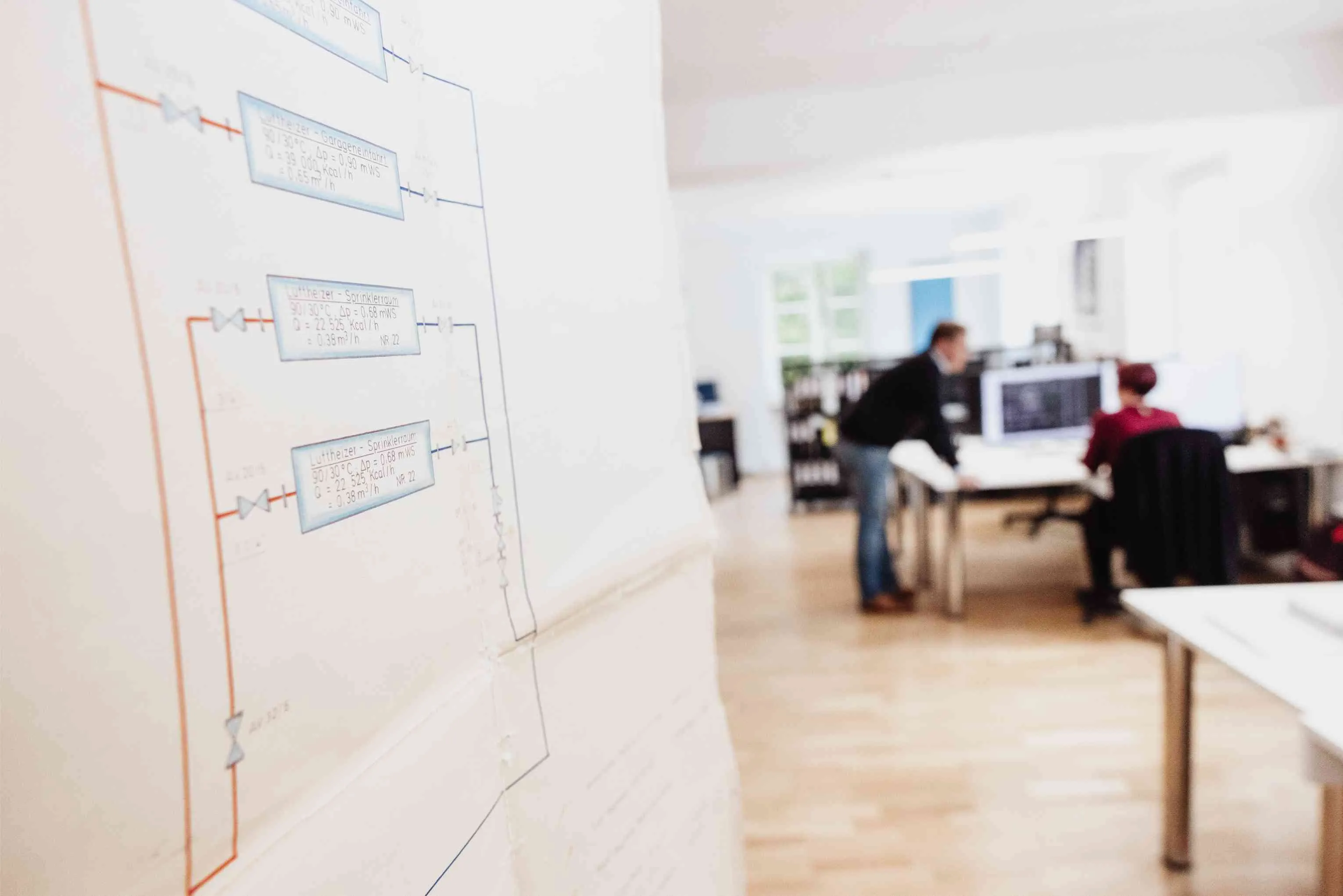 Zwei Personen arbeiten in einem modernen Büro mit Computerbildschirmen, im Vordergrund technische Zeichnungen an einer Wand.