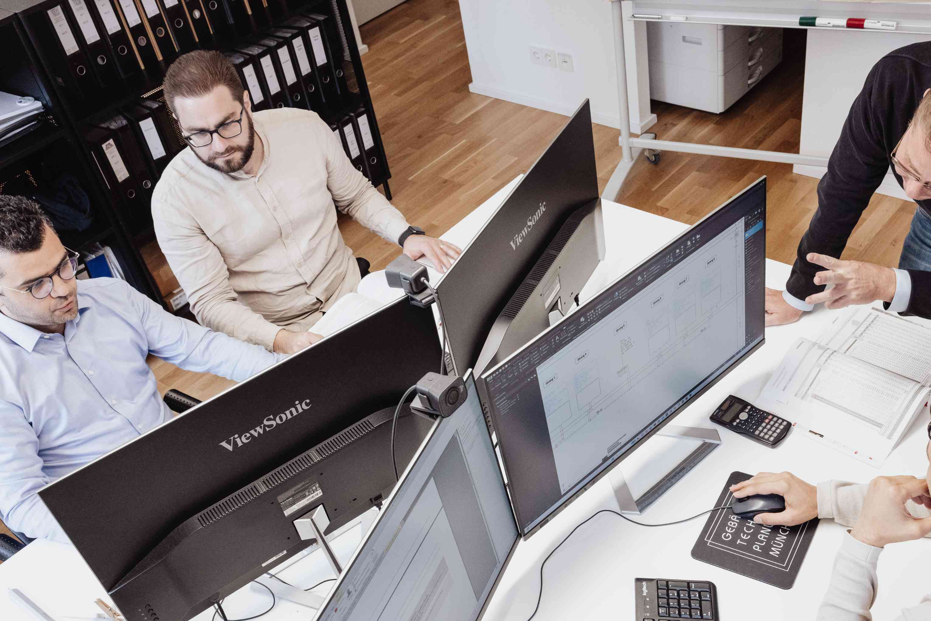 Vier Personen arbeiten gemeinsam an Computern mit mehreren Monitoren in einem Büro mit Ordnerregalen und Whiteboard im Hintergrund.