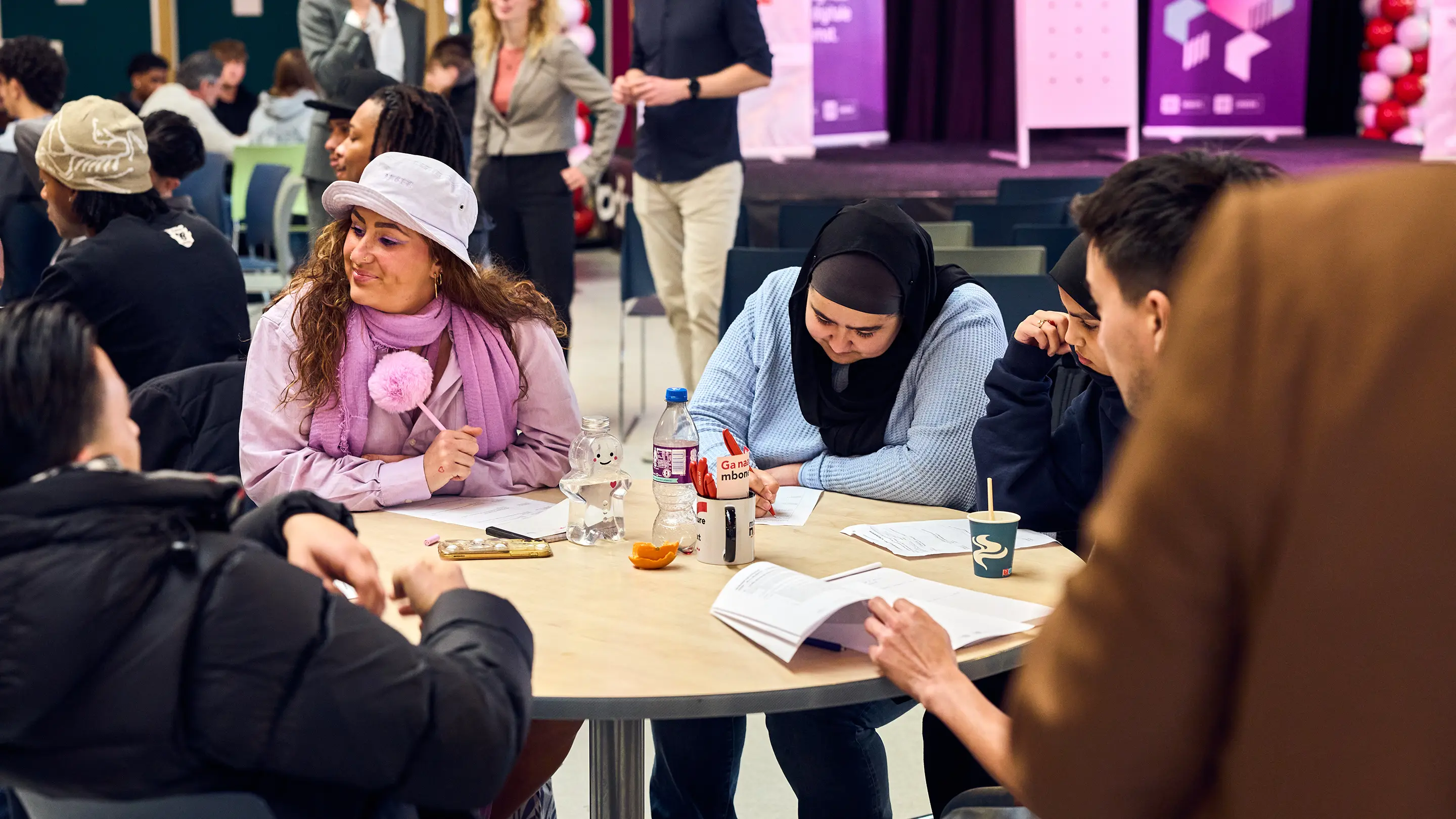 Group of diverse young adults sitting around a round table, working on paperwork and studying together in a casual indoor setting.