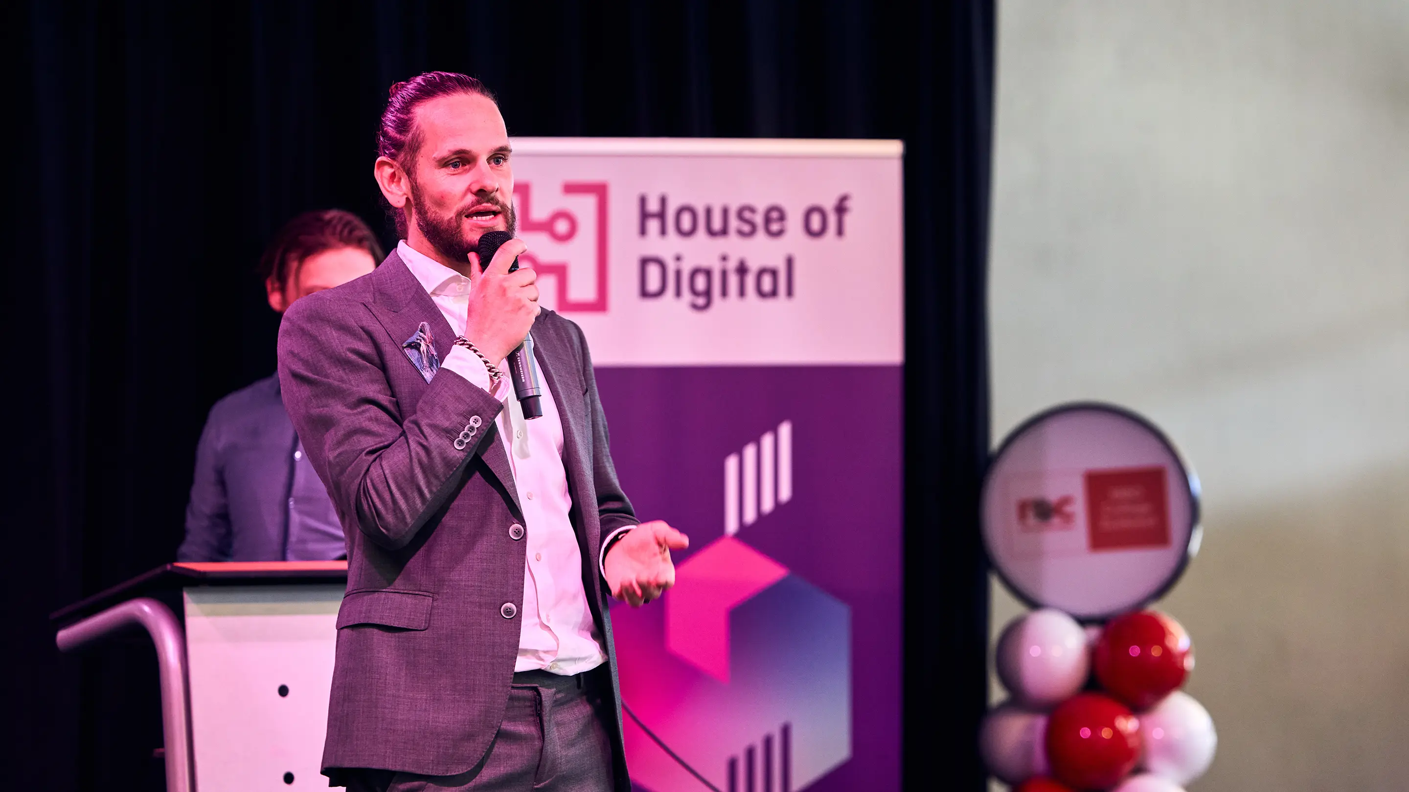 Man in a gray suit speaking into a microphone at a House of Digital event with a banner in the background.