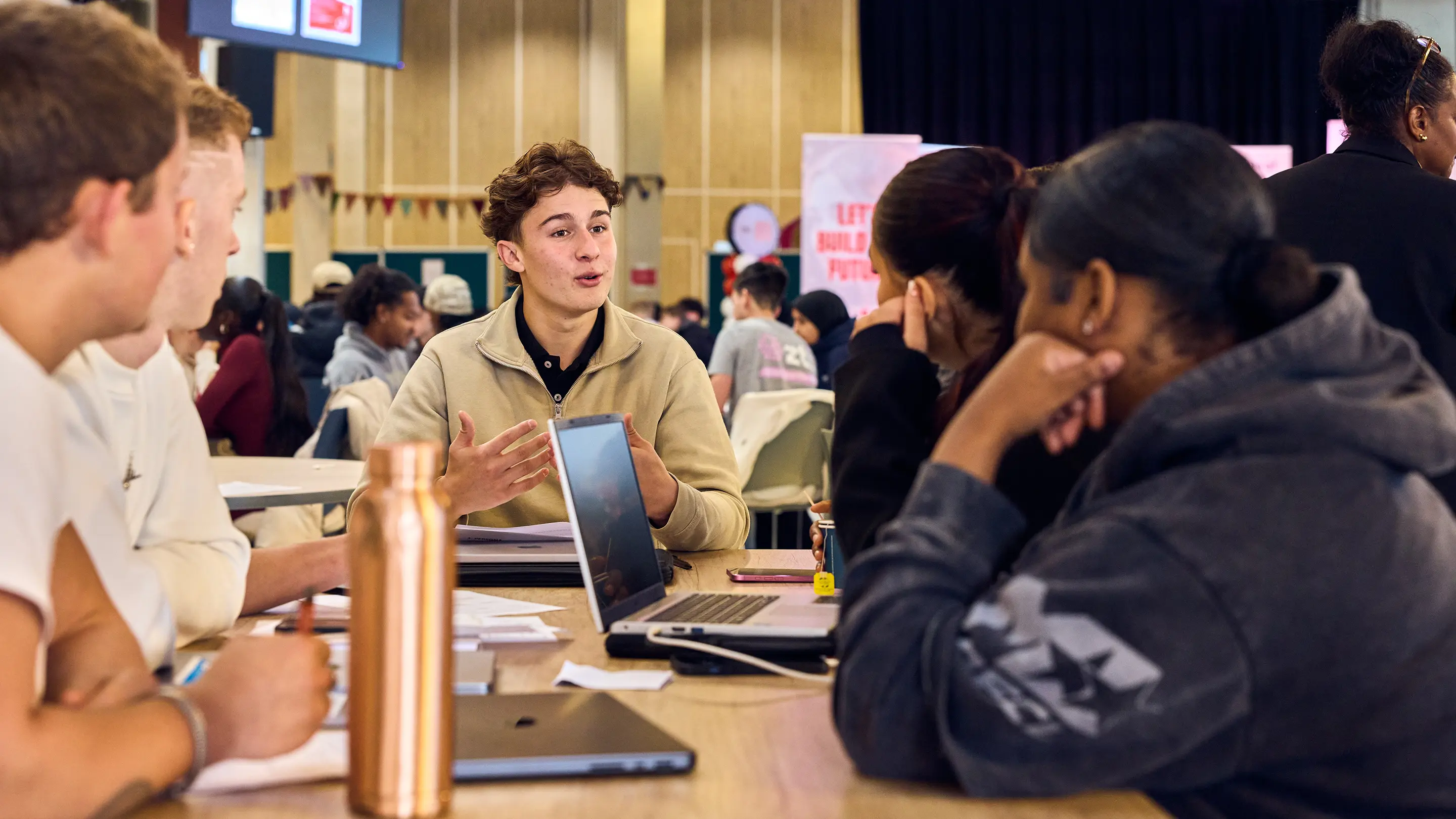 A group of diverse young people sitting around a table engaged in a discussion with laptops and notebooks.