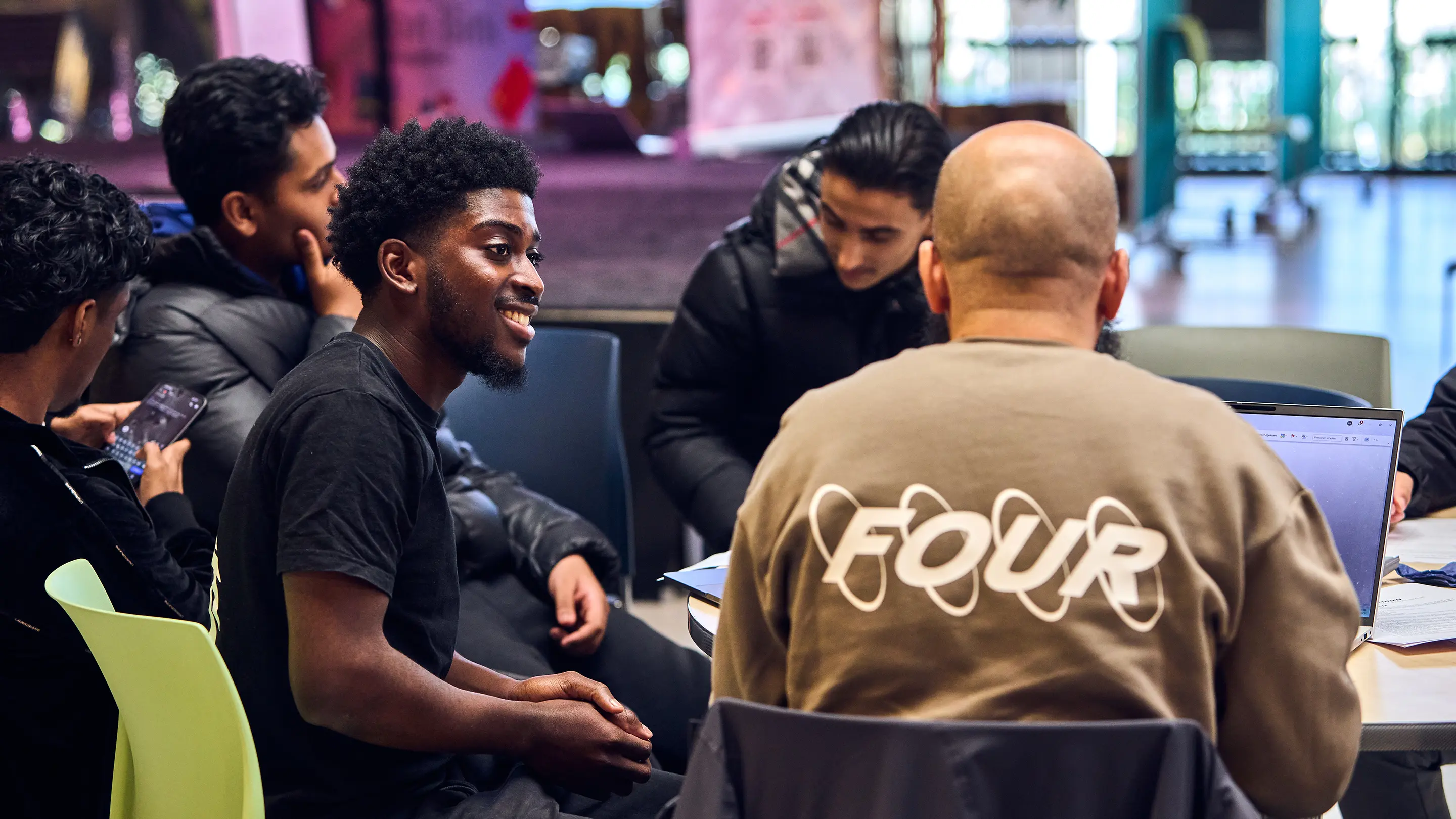 Group of five young men sitting around a table engaged in conversation, with one smiling and another using a laptop.