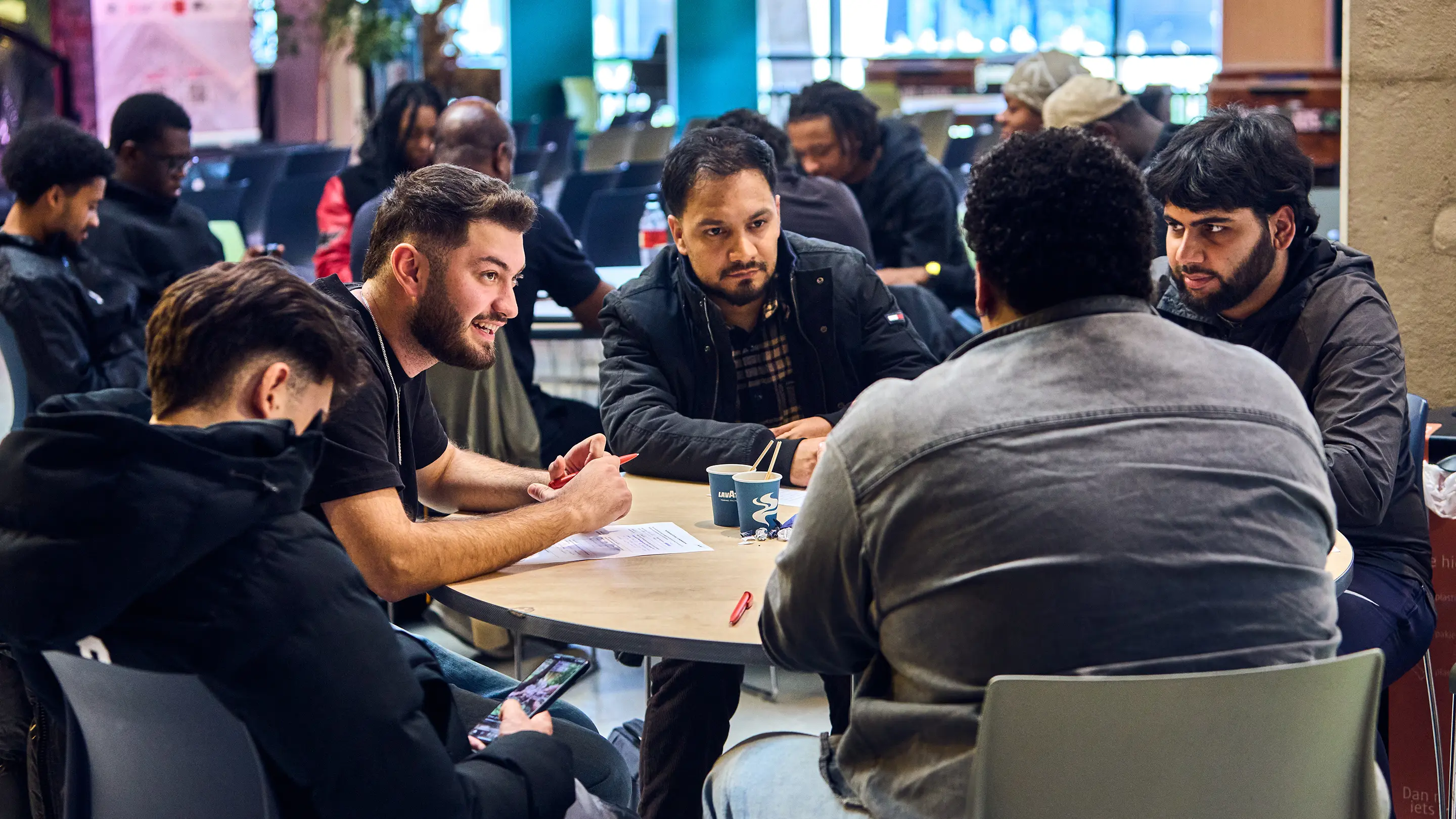 A group of diverse young men sitting around a table engaged in conversation and writing on papers in a casual indoor setting.
