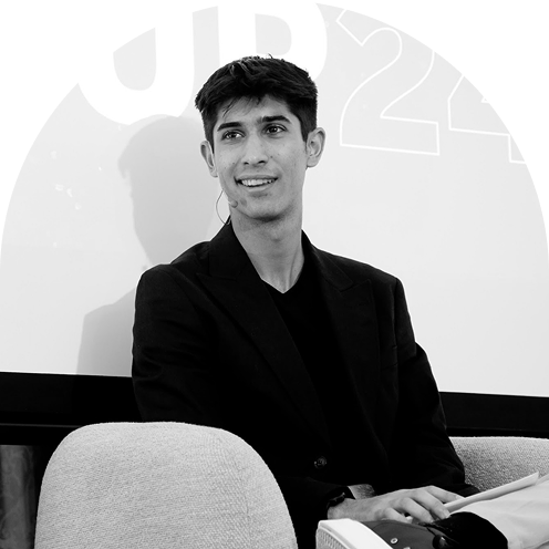 Young man in black blazer sitting on a chair, smiling and holding a notebook and pen.