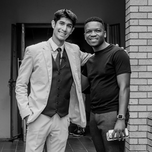 Two men smiling and posing together, one in a suit and the other in a black shirt holding a phone.