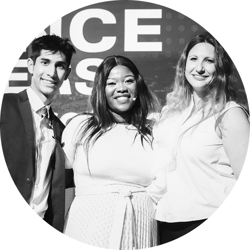 Three smiling people posing together in front of a sign, one man and two women in professional attire.