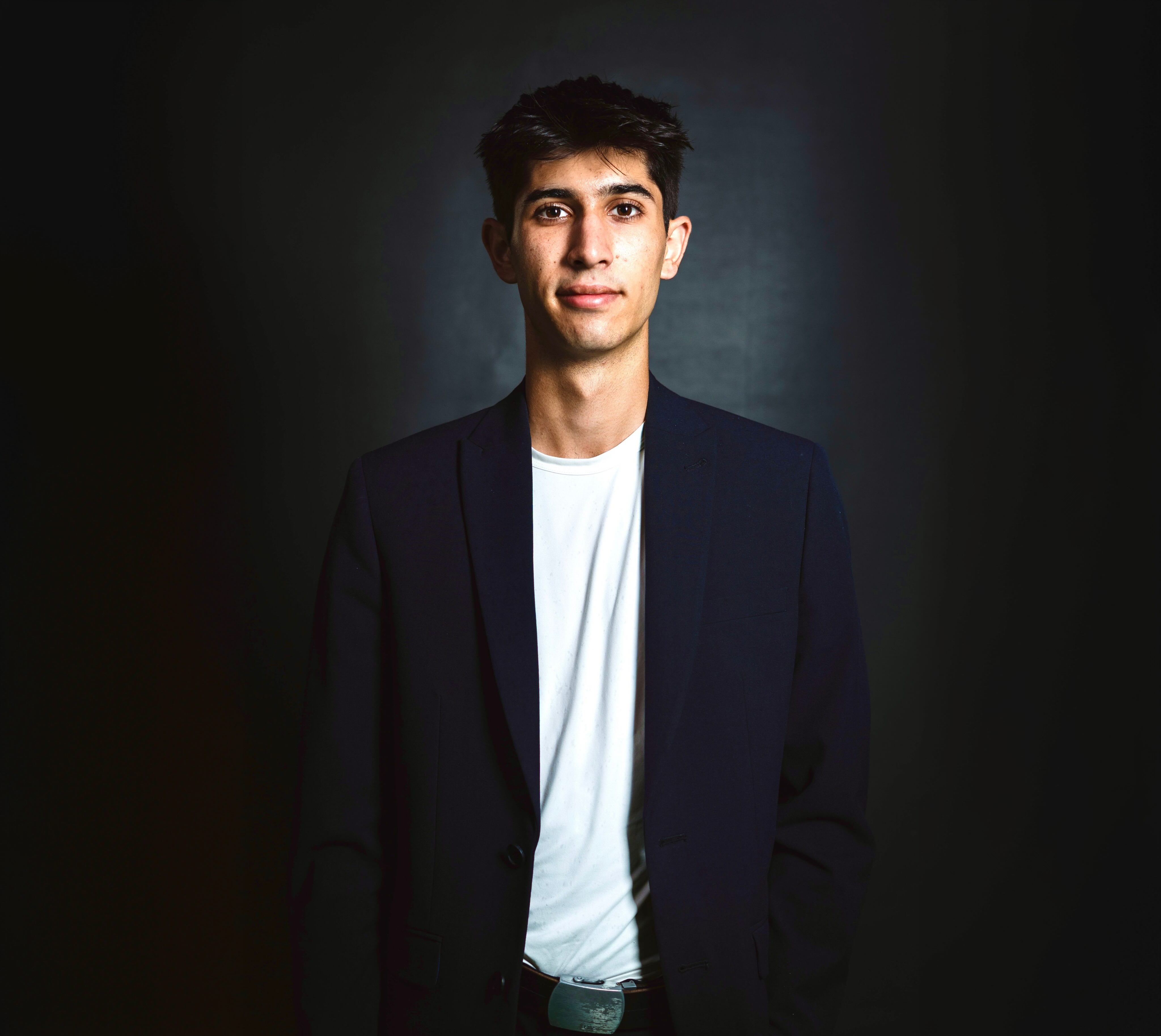 Young man in a white shirt and dark blazer standing against a dark background.