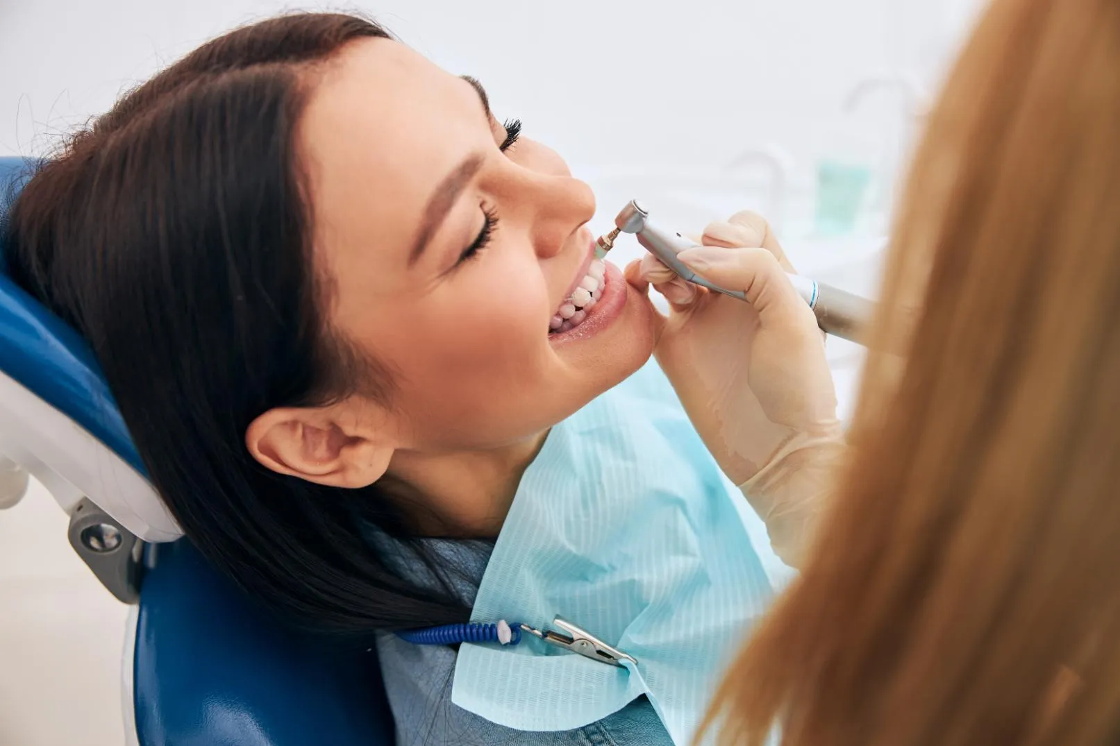 Woman smiling with eyes closed while a dentist cleans her teeth using a dental tool.