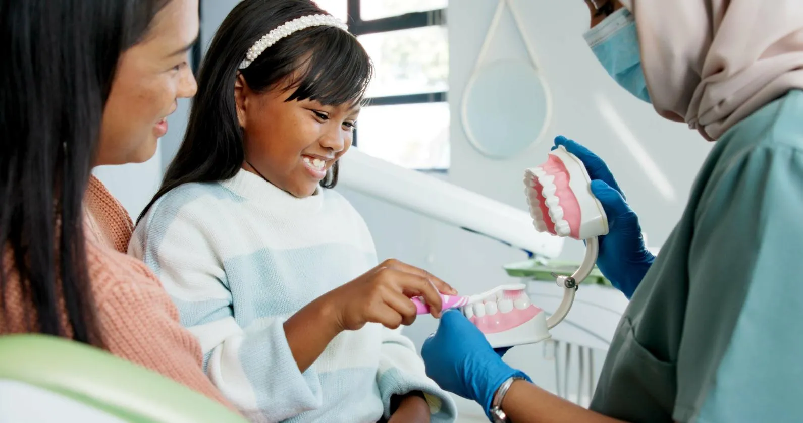 Child learning to brush teeth on a dental model with a toothbrush, guided by a masked dentist wearing gloves, while the mother watches.