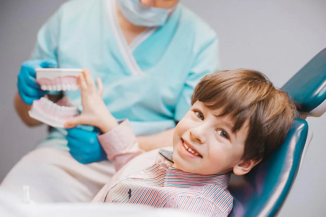 Smiling young boy sitting in a dental chair reaching for a dental model held by a dentist in scrubs and gloves.