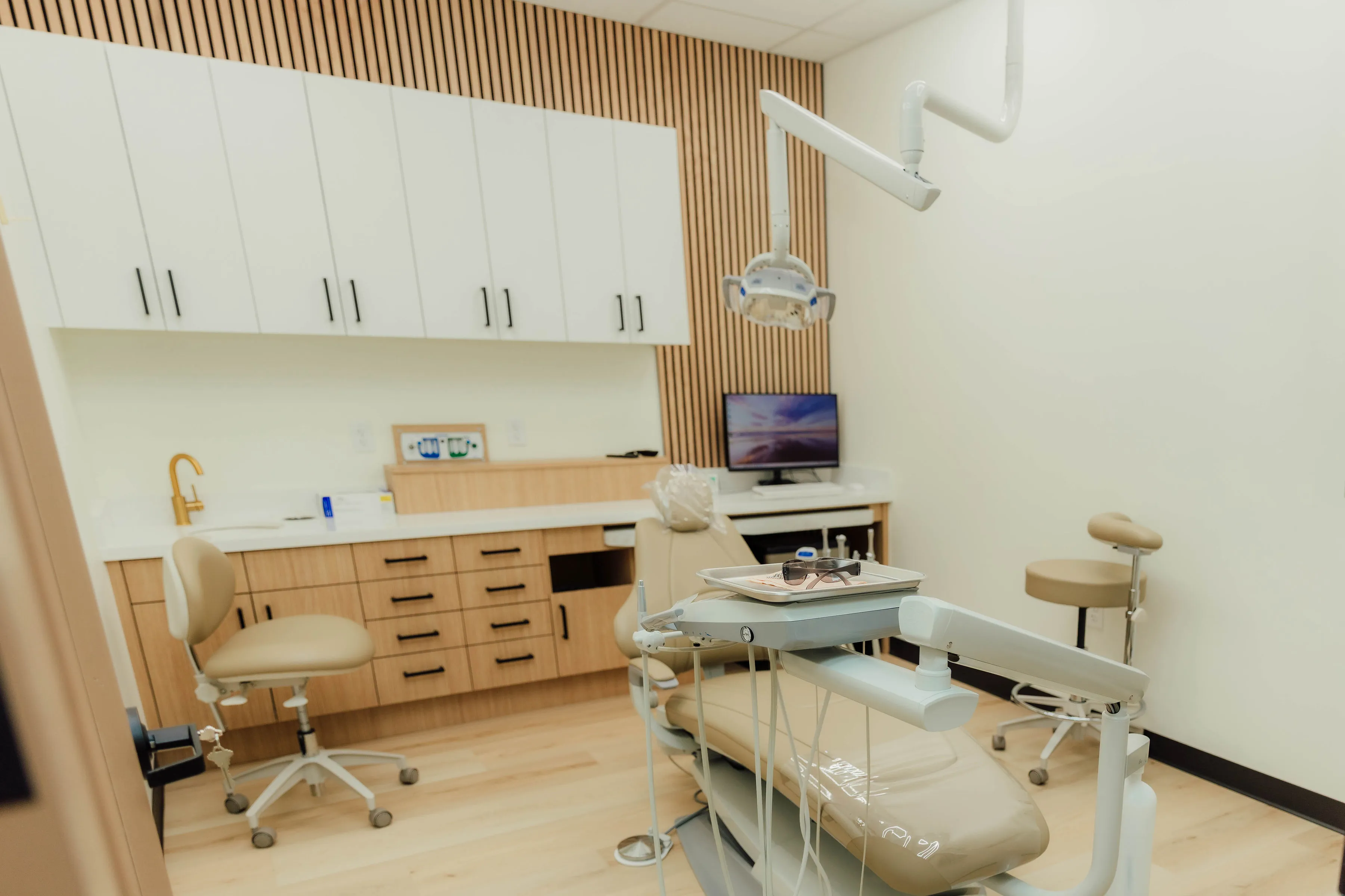 Modern dental clinic room with beige dental chair, overhead light, tray with protective glasses, cabinets, and a computer monitor in clayton dental studio Humble, Tx