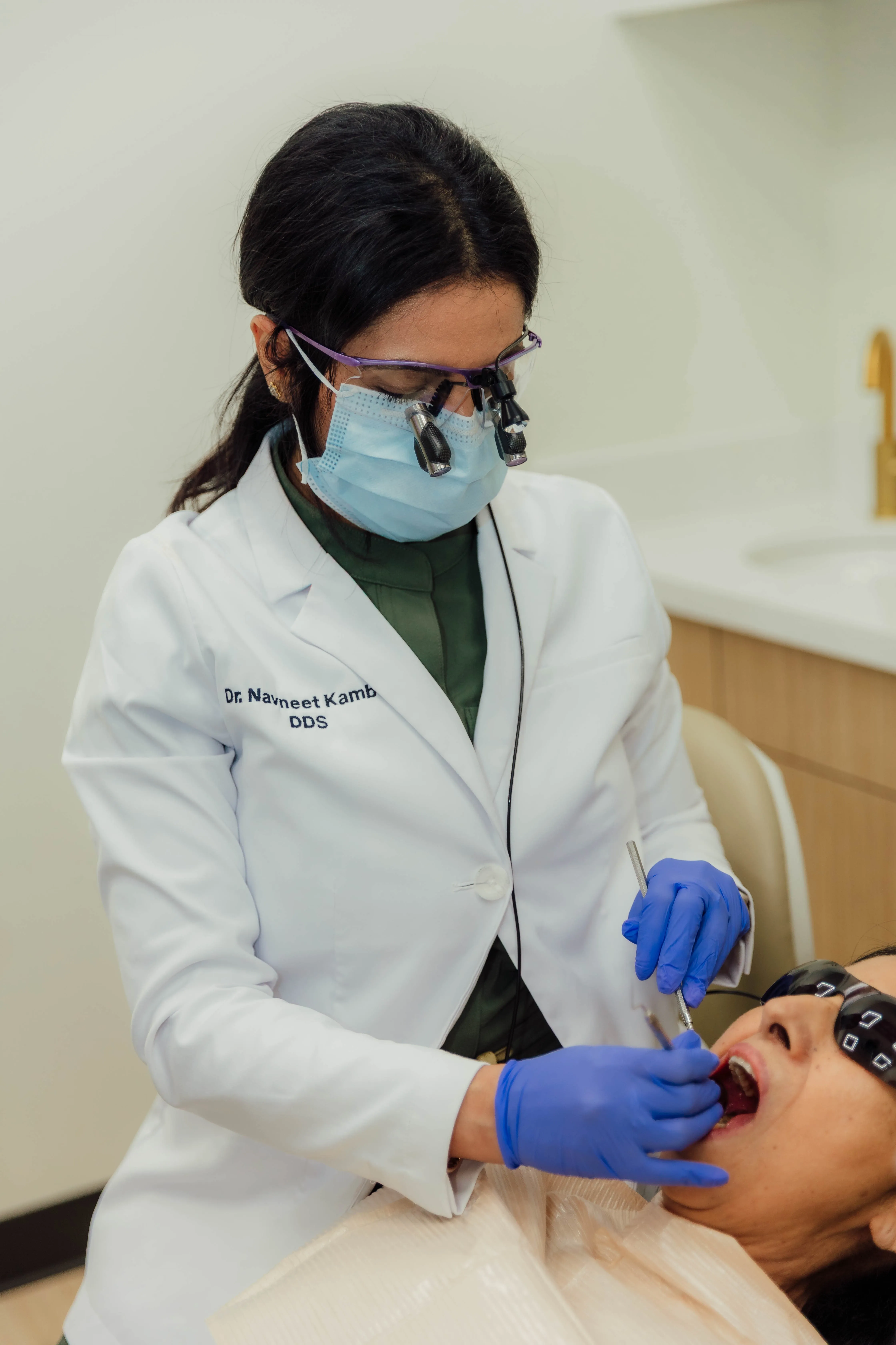 Dentist wearing a mask, magnifying glasses, and gloves examines a patient's mouth with dental tools.
