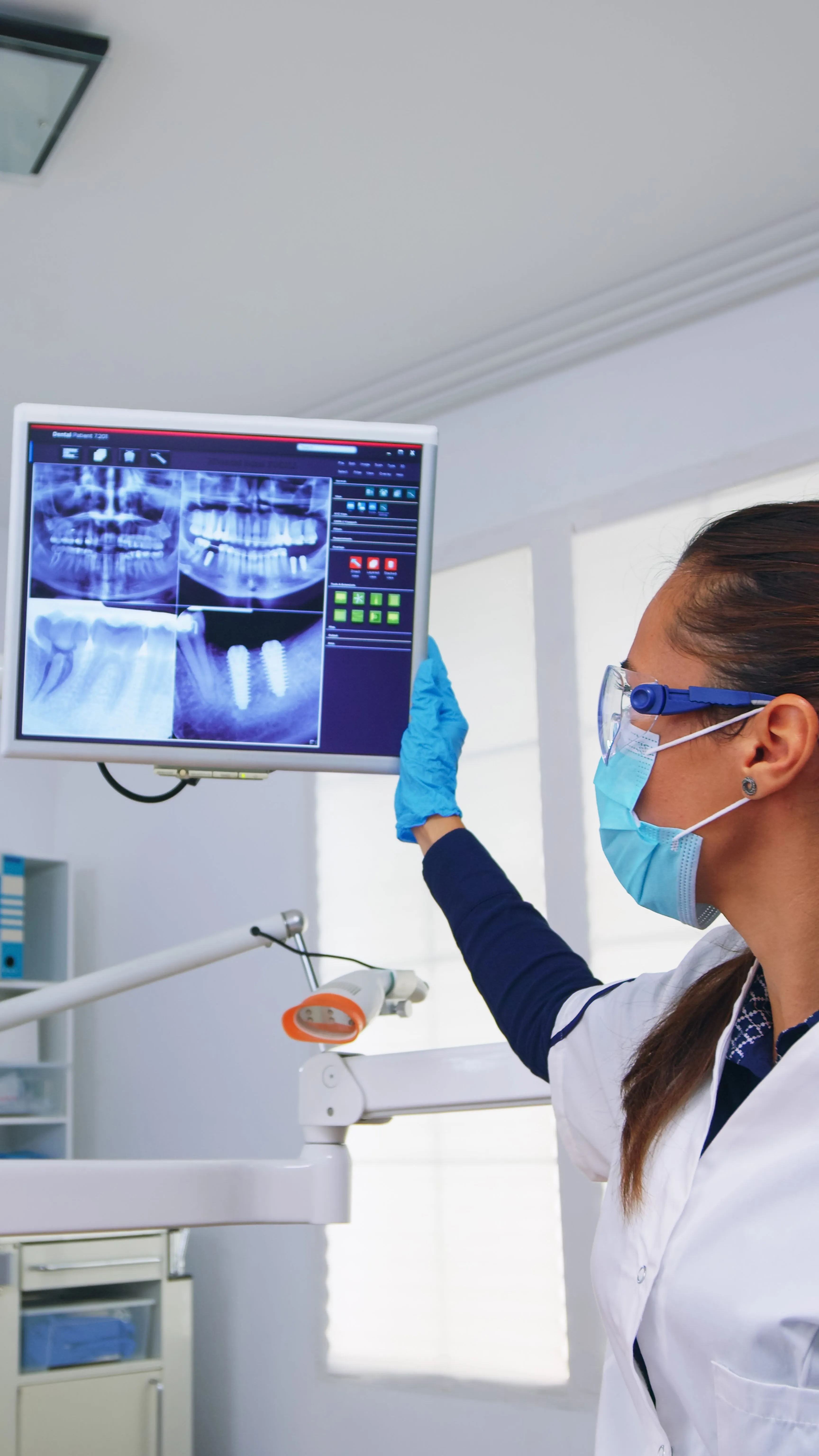Dentist wearing a mask and protective glasses holding a screen displaying four dental x-rays in a clinic.