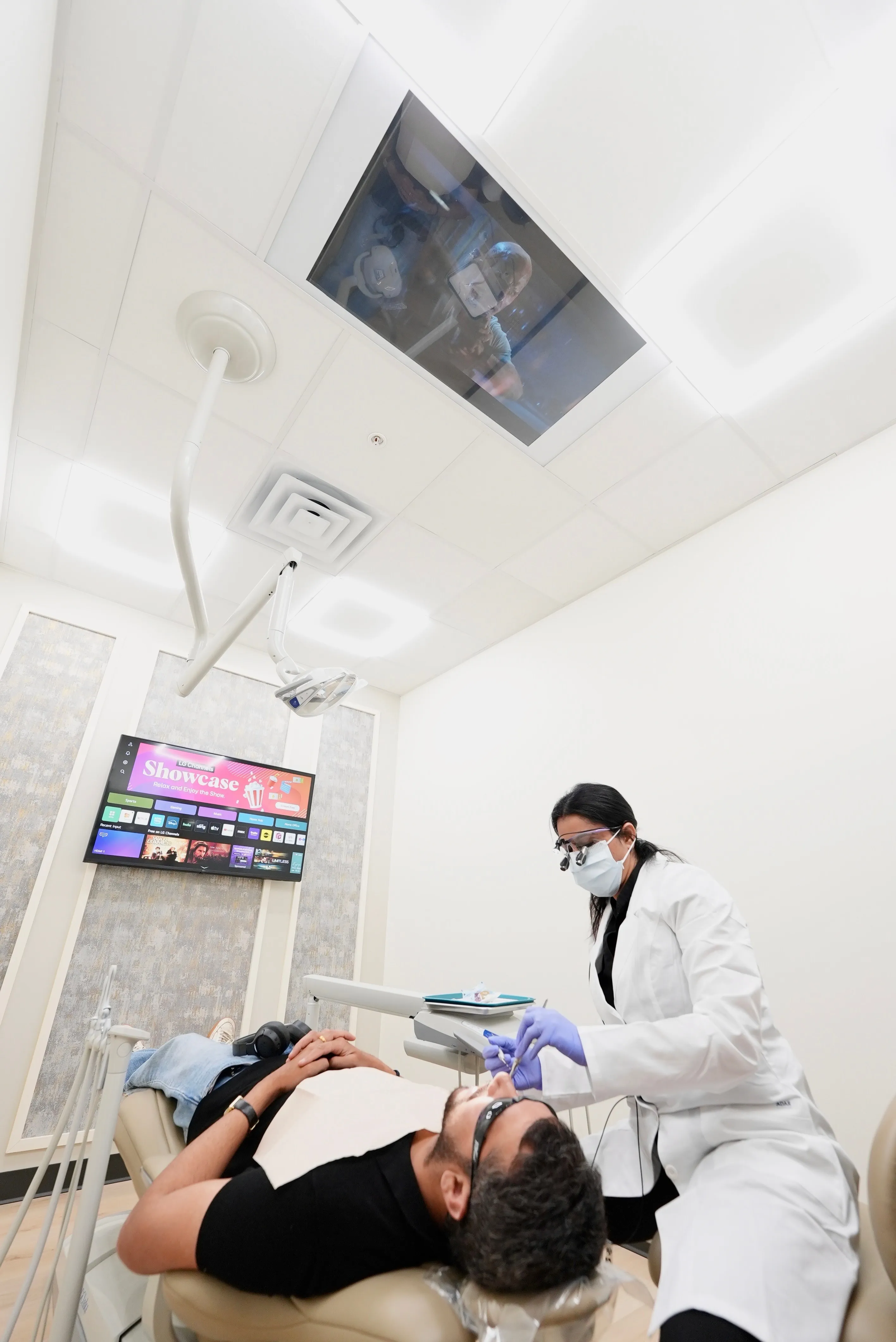 Dentist in white coat and mask treating a patient lying on a dental chair with a television screen on the wall.