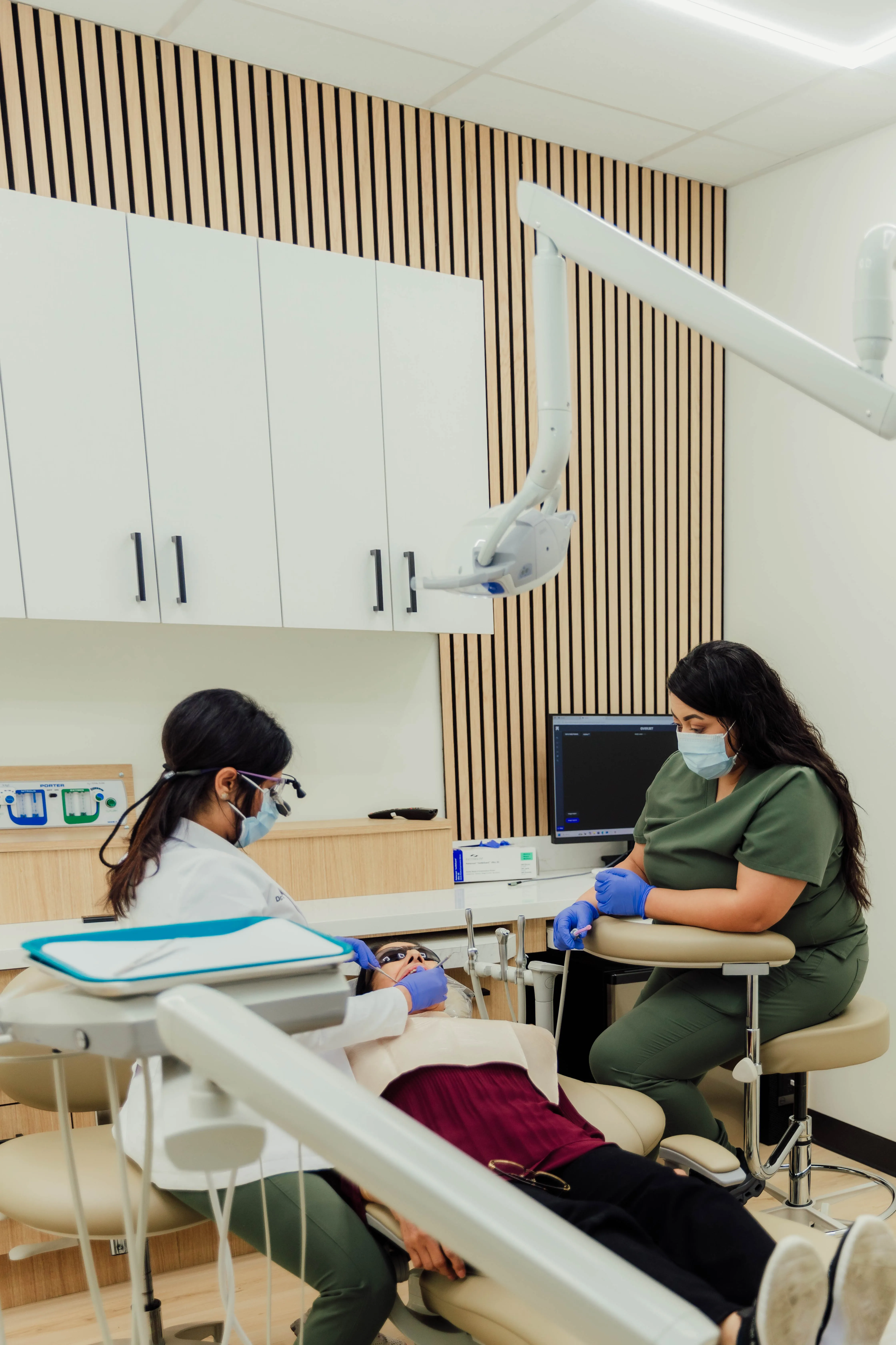 Dental hygienist wearing a mask and gloves examining a patient lying in a dental chair, with another staff member observing. Both are in Clayton dental studio.